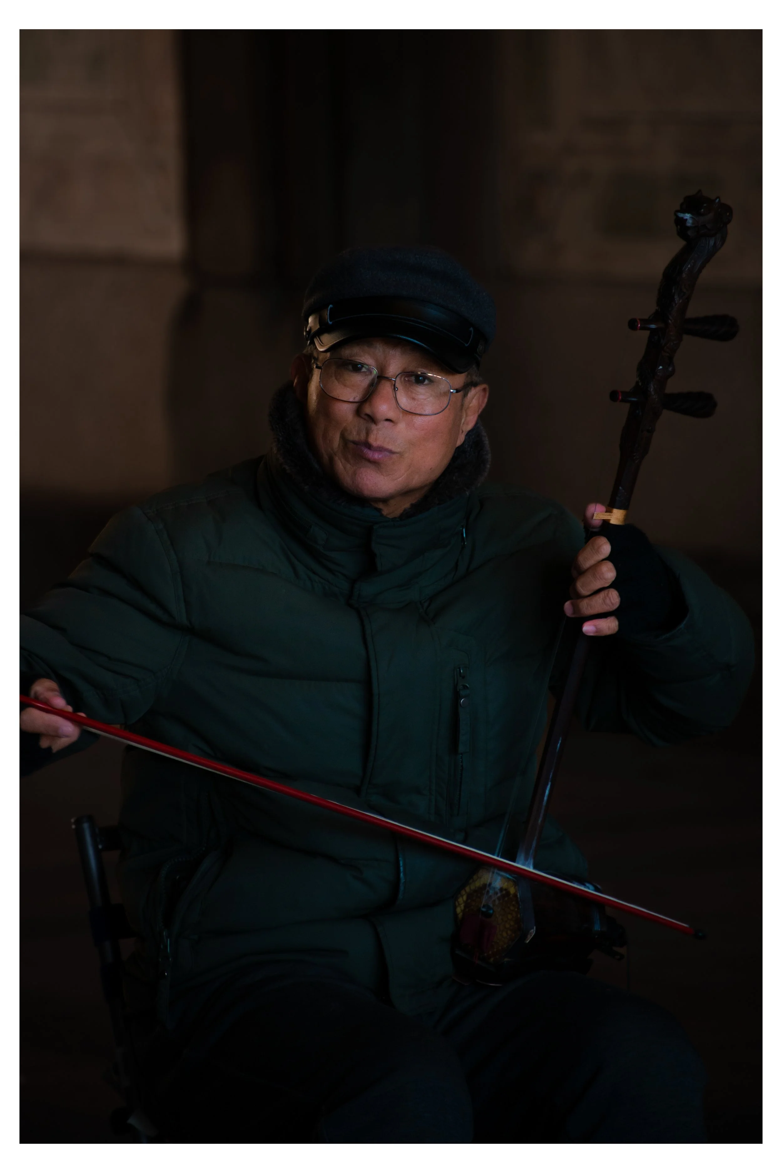 A man wearing glasses, a dark jacket, a black cap, and gloves, sitting on a chair in a dimly lit indoor setting, holding a walking stick.