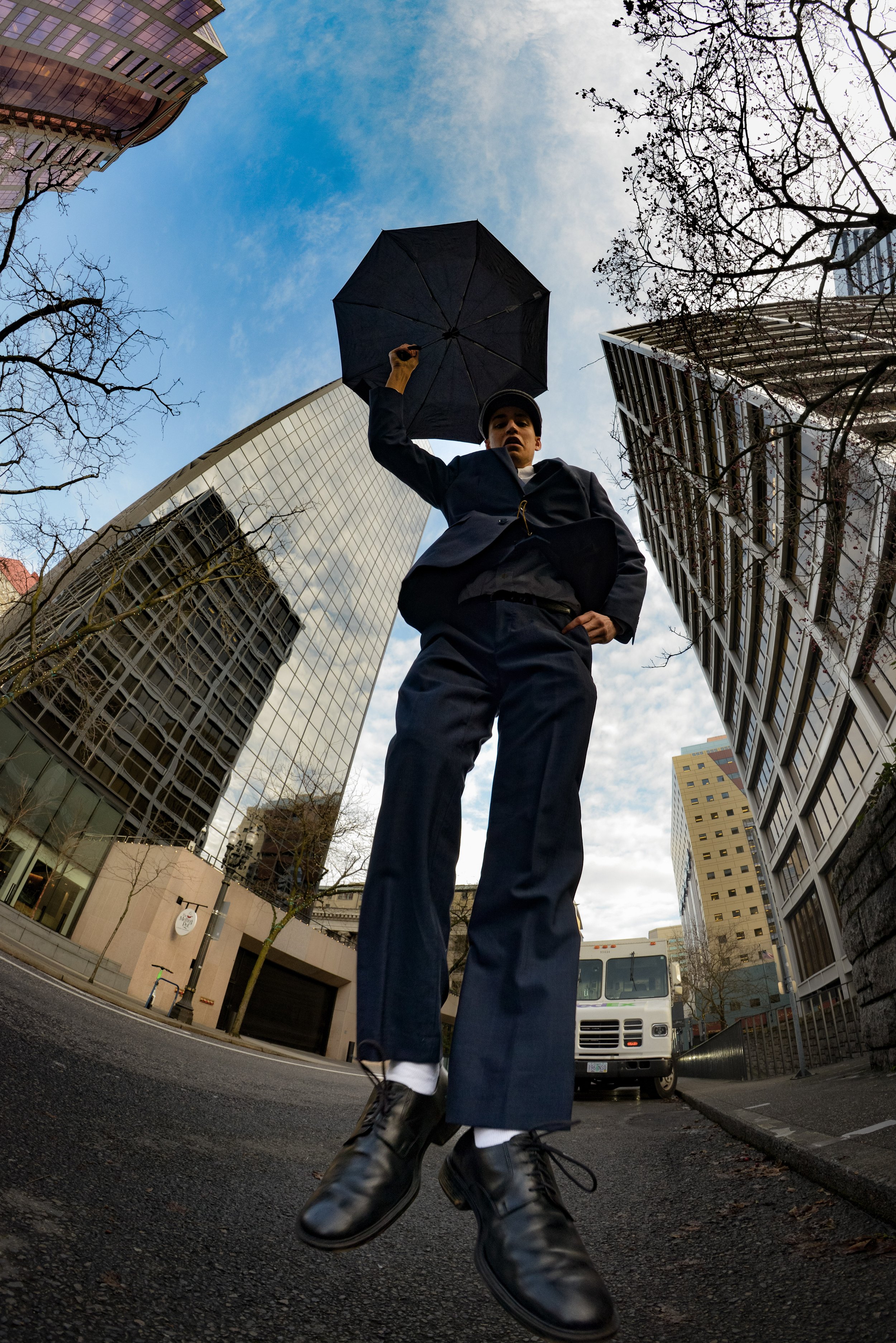 A person in business attire holding a black umbrella, standing on a city street surrounded by tall buildings, with the view looking up from below.