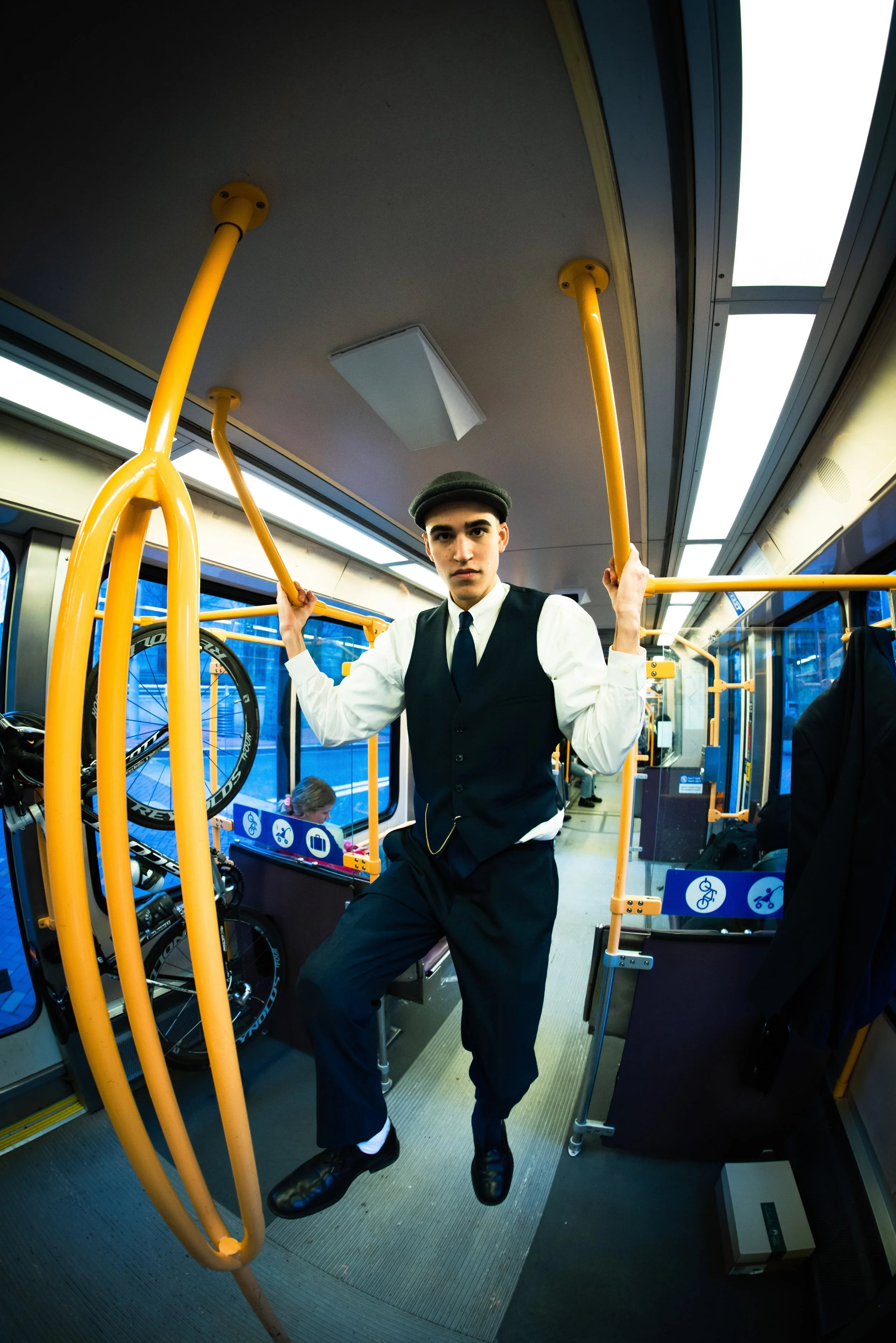 A young man dressed in a vintage train conductor uniform with a gray cap, white shirt, black vest, and black trousers, standing inside a bus near the door. He is holding onto the yellow handrails and looking directly at the camera. The bus interior f