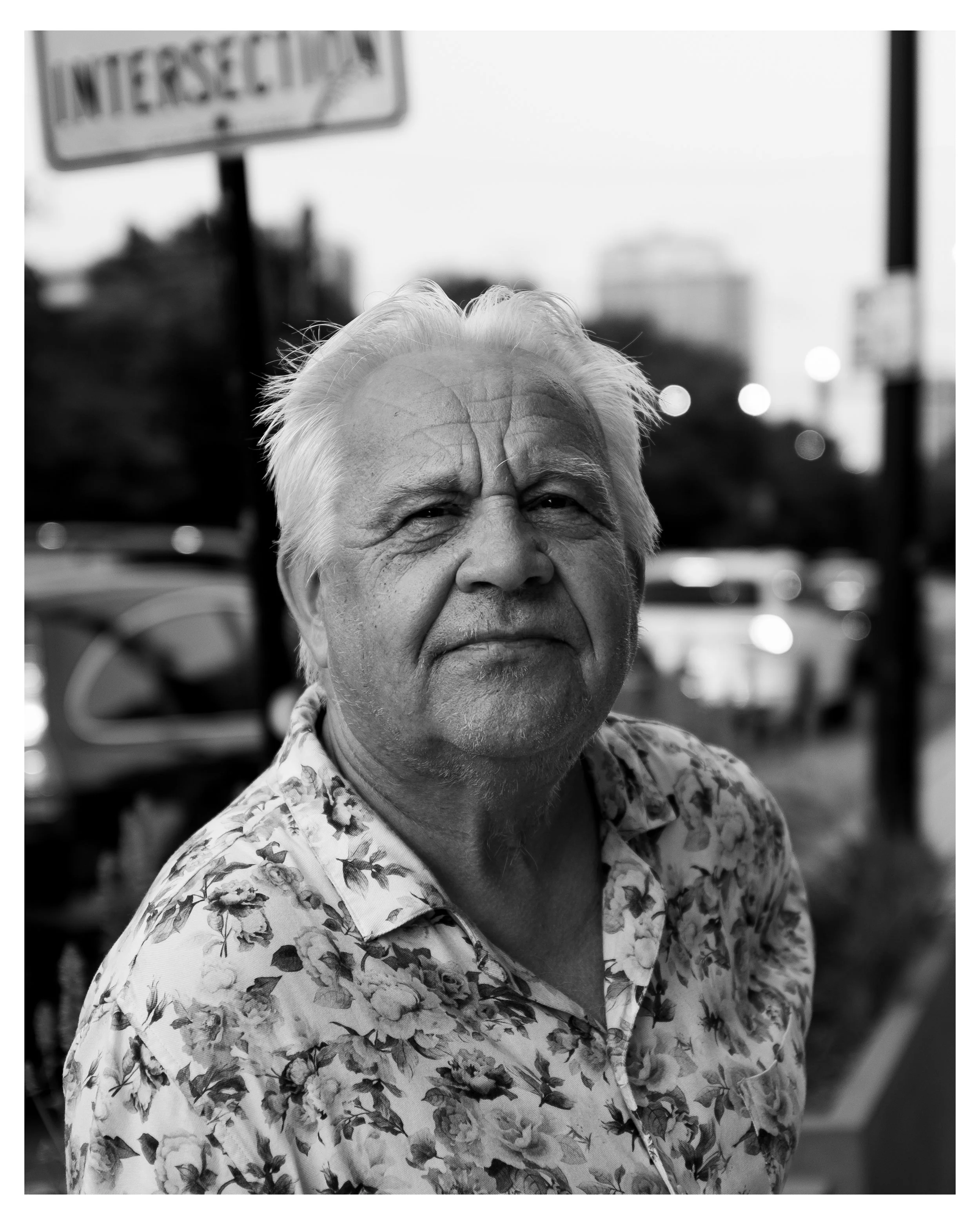 Close-up black and white photo of an elderly man with white hair and wrinkles, wearing a patterned shirt with floral designs, outdoors with blurred cars and city buildings in the background.