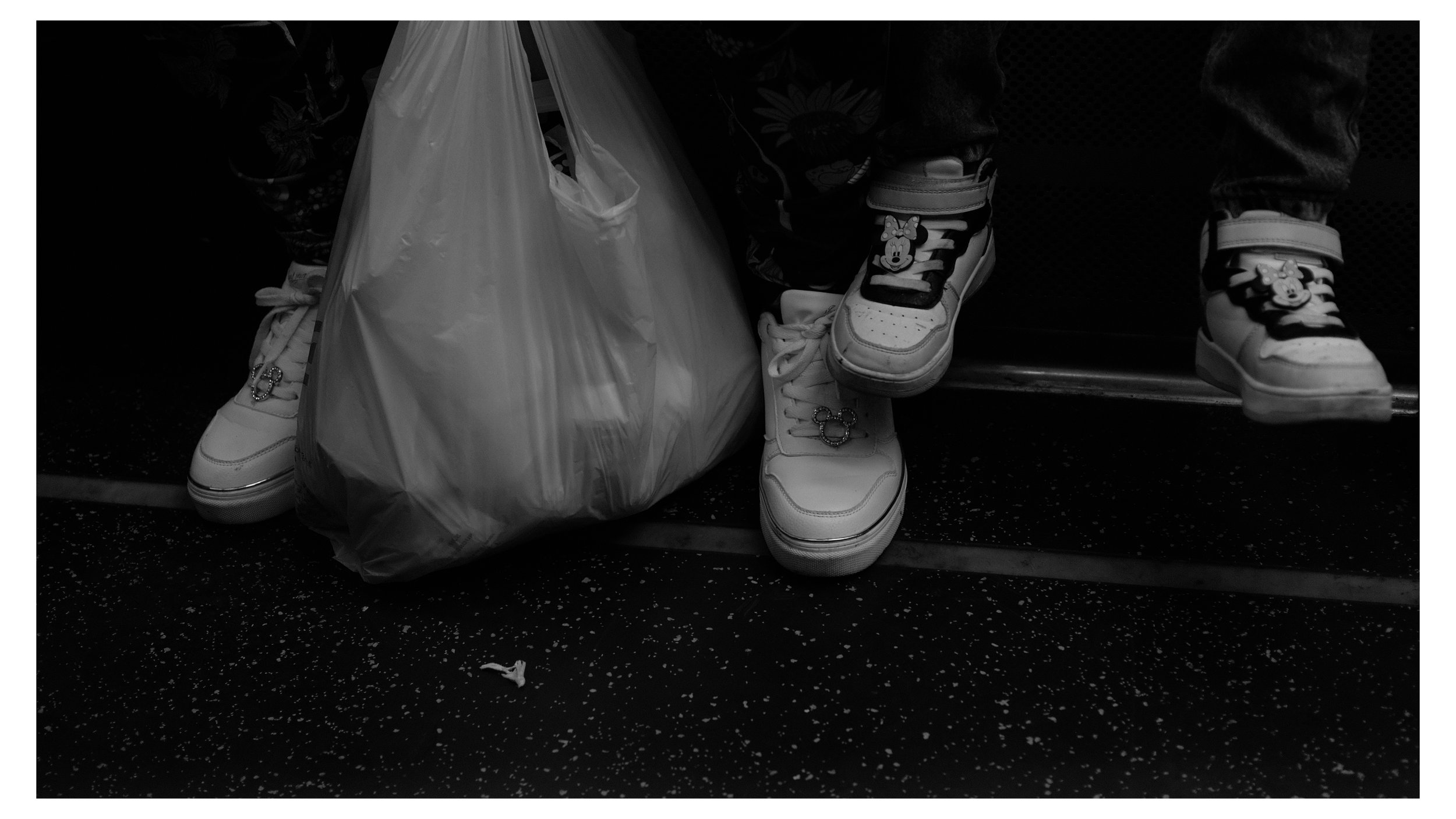 Close-up of a person's feet wearing Minnie Mouse-themed sneakers with velcro straps, sitting on a bench with a plastic bag hanging nearby.