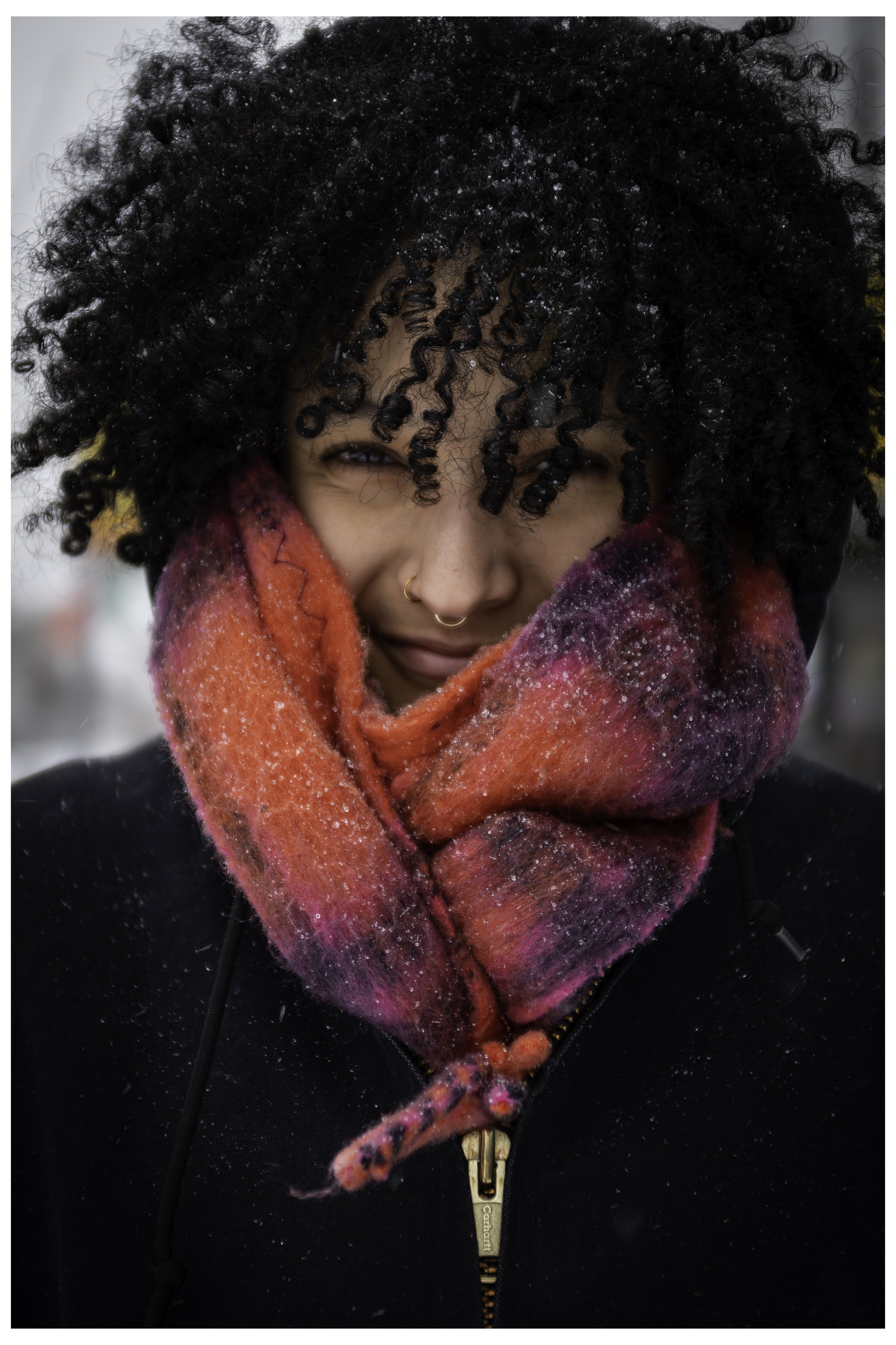 Close-up of a woman smiling, wearing a vibrant pink and purple scarf with snowflakes, black jacket with a gold zipper, and a black coat, outdoors in a snowy environment.