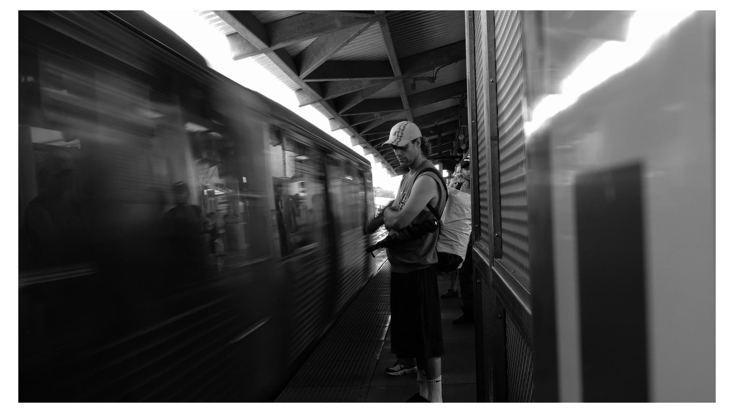 A man in casual clothing, including a cap, standing on a train platform and waiting to board the train, with another person behind him, all in black and white.