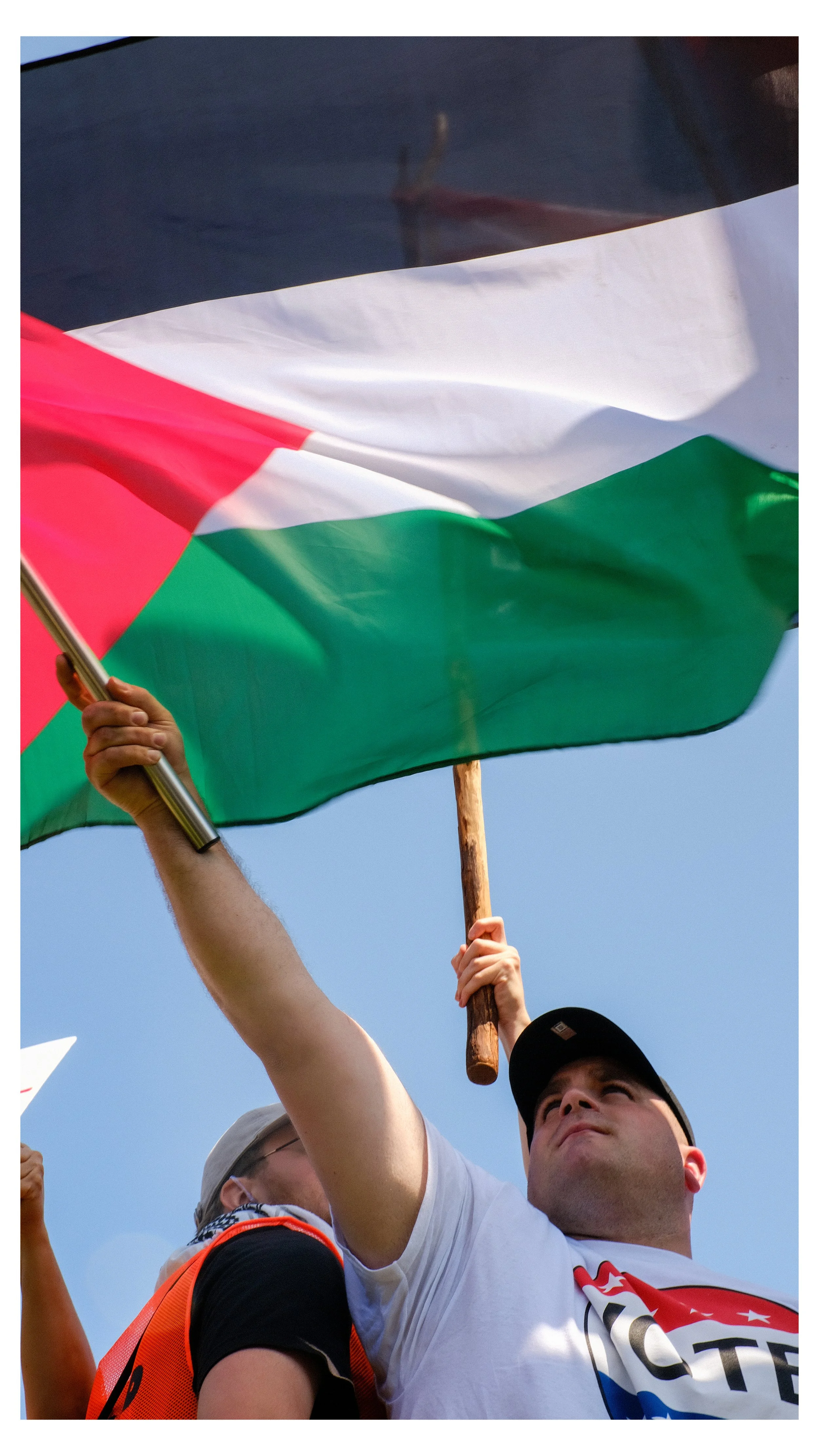 Two people holding up a Palestinian flag during a demonstration, with a clear blue sky in the background.