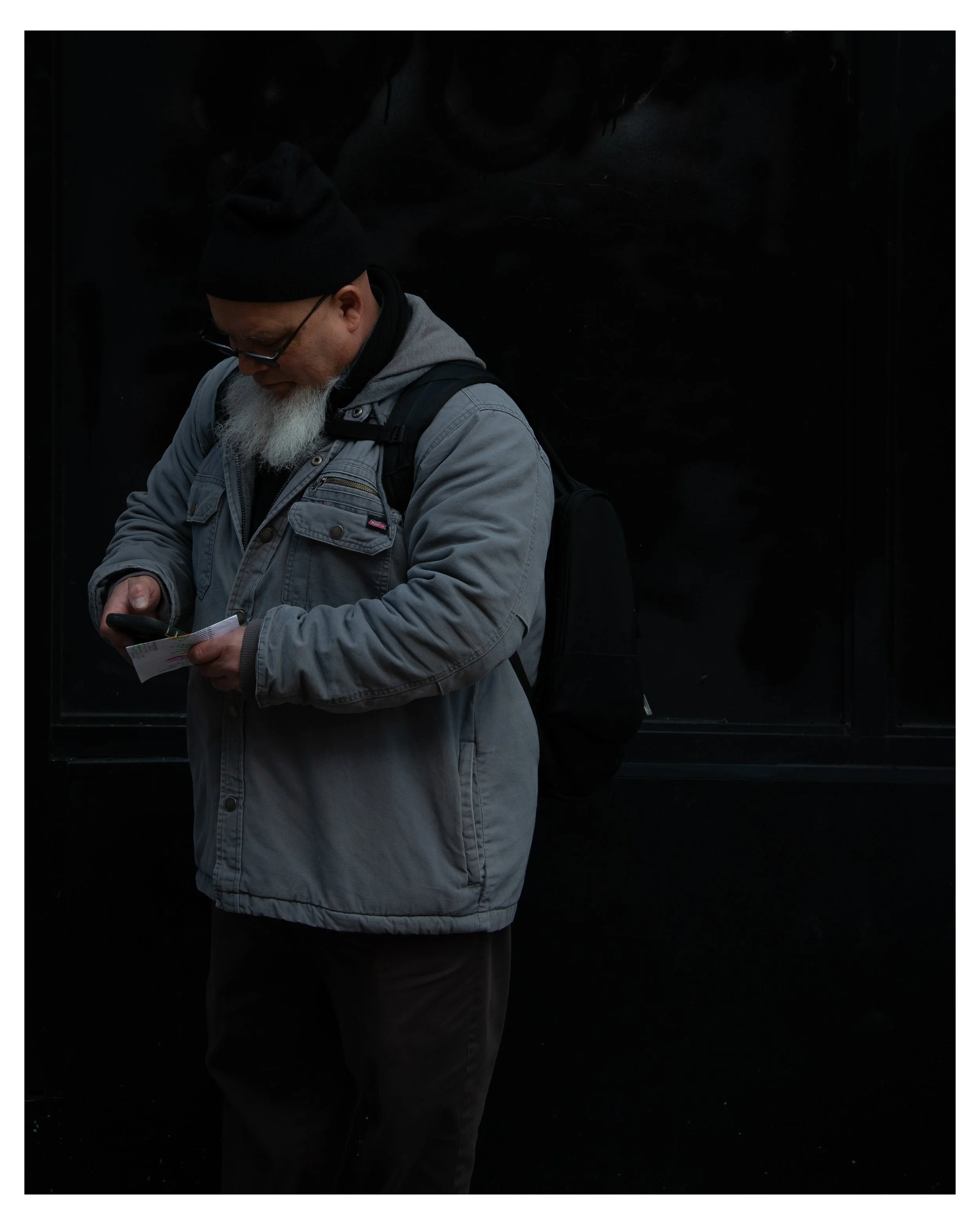 A man with a grey jacket, black beanie, glasses, and a white beard, looking down at his phone, standing against a black background.