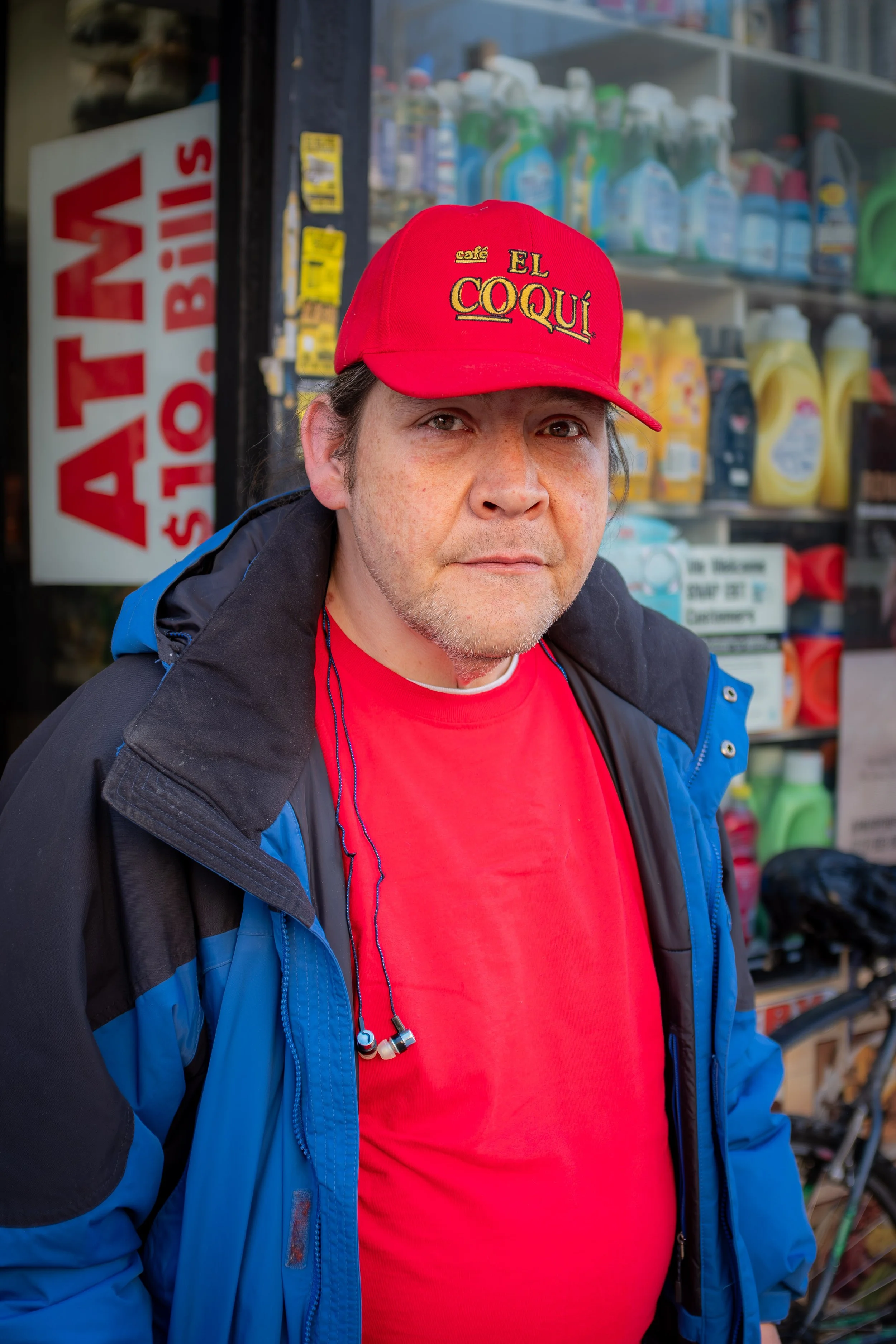A man with a light beard and freckles wearing a red cap with yellow embroidery that says 'El Coquí' and a red shirt. He is standing outside near a store with cleaning supplies in the background.