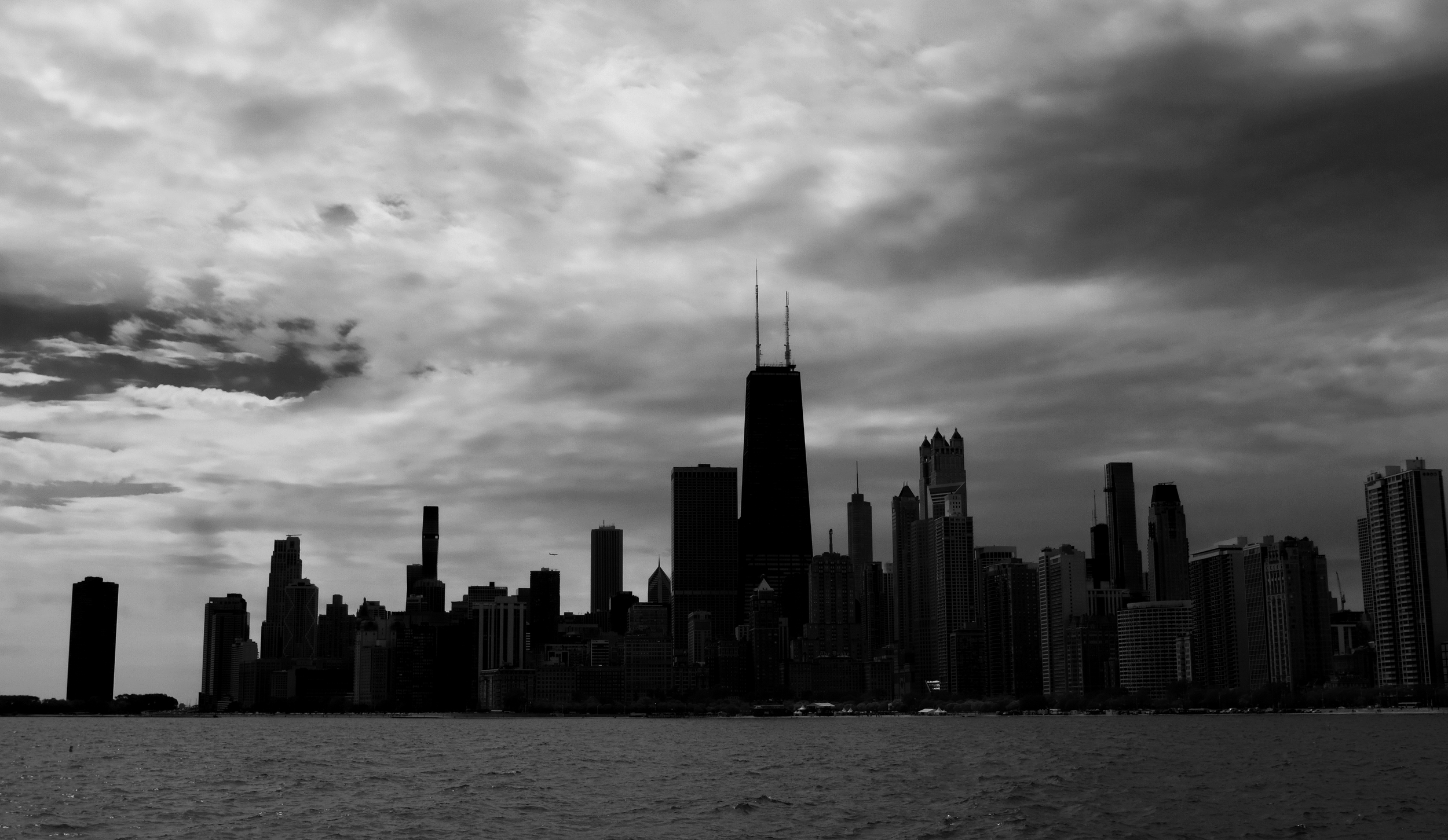 Black and white photo of a city skyline with tall buildings, including a prominent skyscraper with two antennas, viewed from across a body of water under a cloudy sky.
