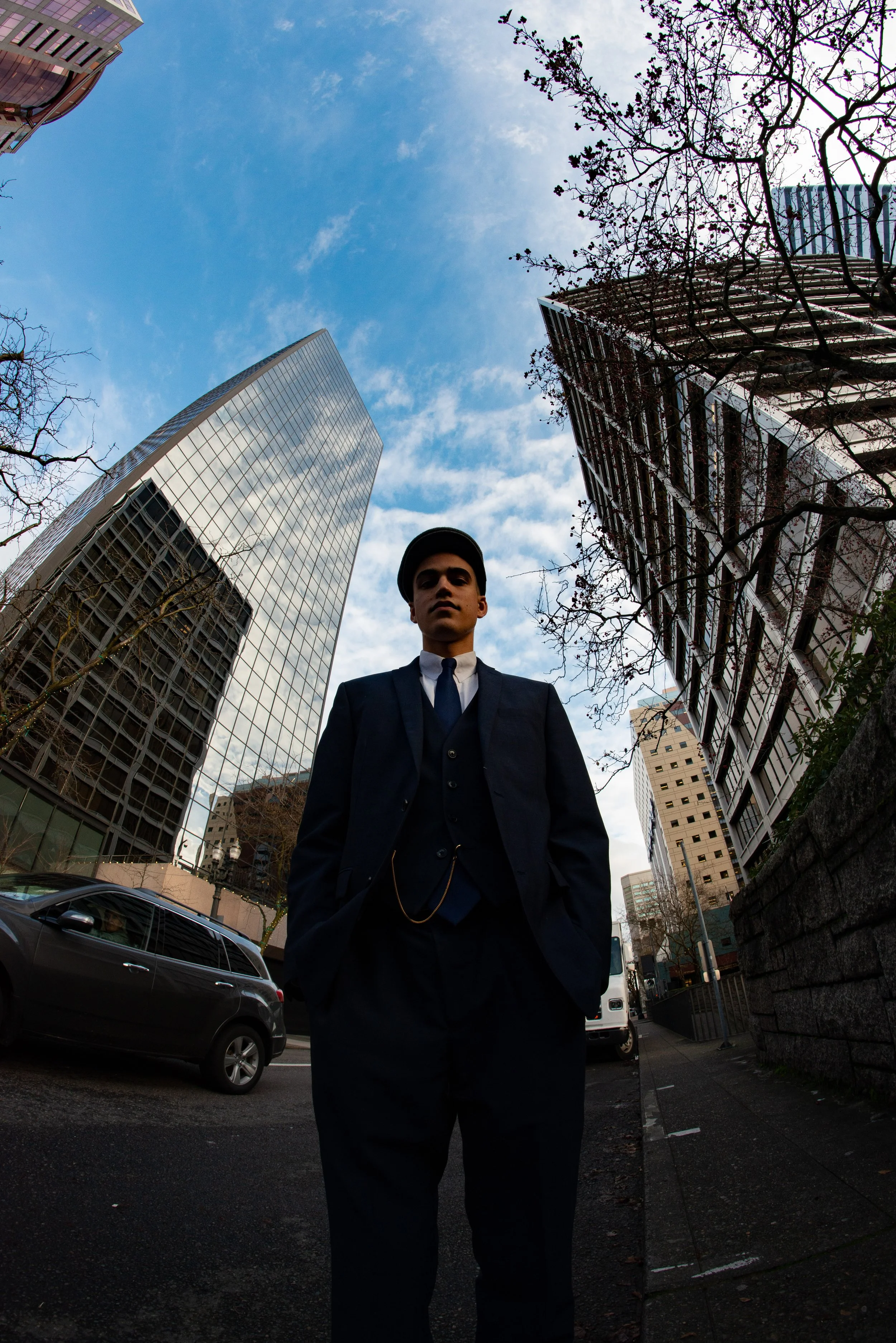 A young man in a dark suit and tie stands on a city street, with tall modern buildings behind him and a clear blue sky above.
