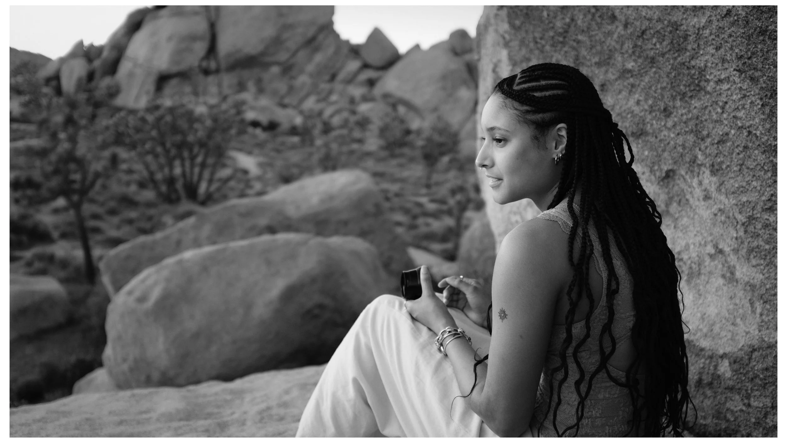 A woman with braided hair sitting against a large rock in an outdoor rocky landscape, holding a camera in her hands and looking into the distance.