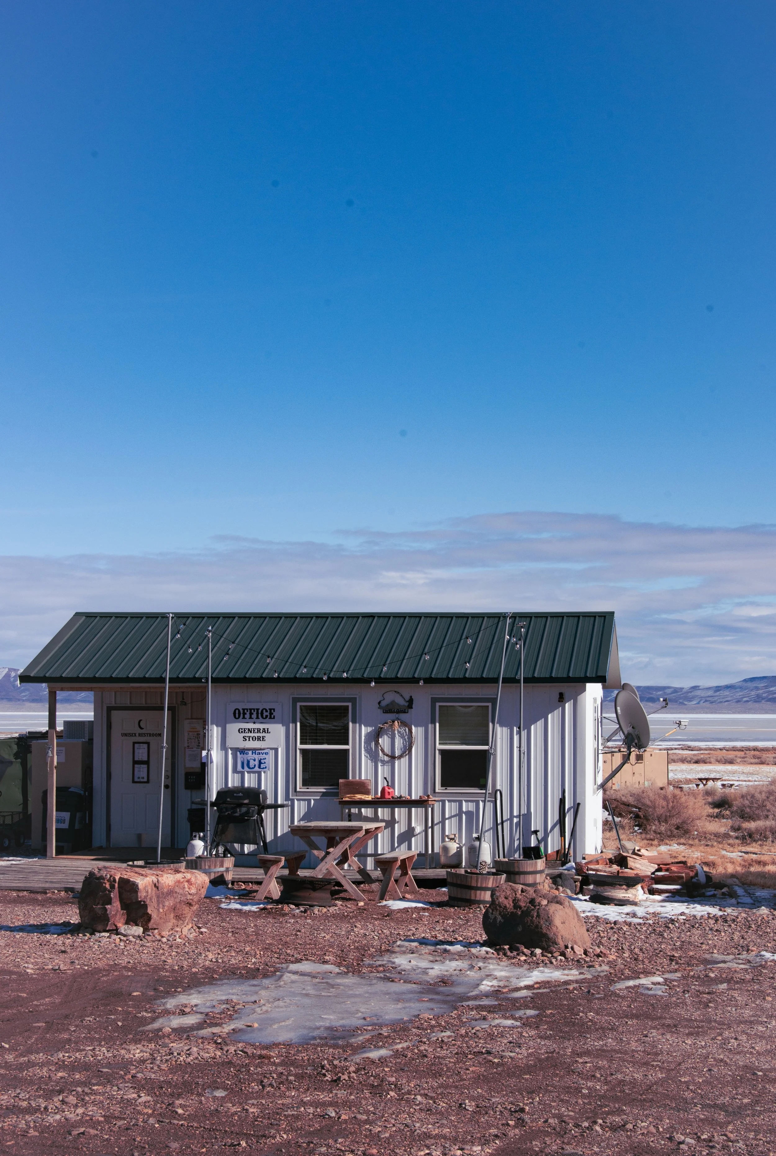 A small white building with a green metal roof, labeled 'Office' and 'General Store,' with outdoor seating, a small sign that says 'We Have Ice,' and a satellite dish, against a landscape of mountains under a blue sky with some clouds.