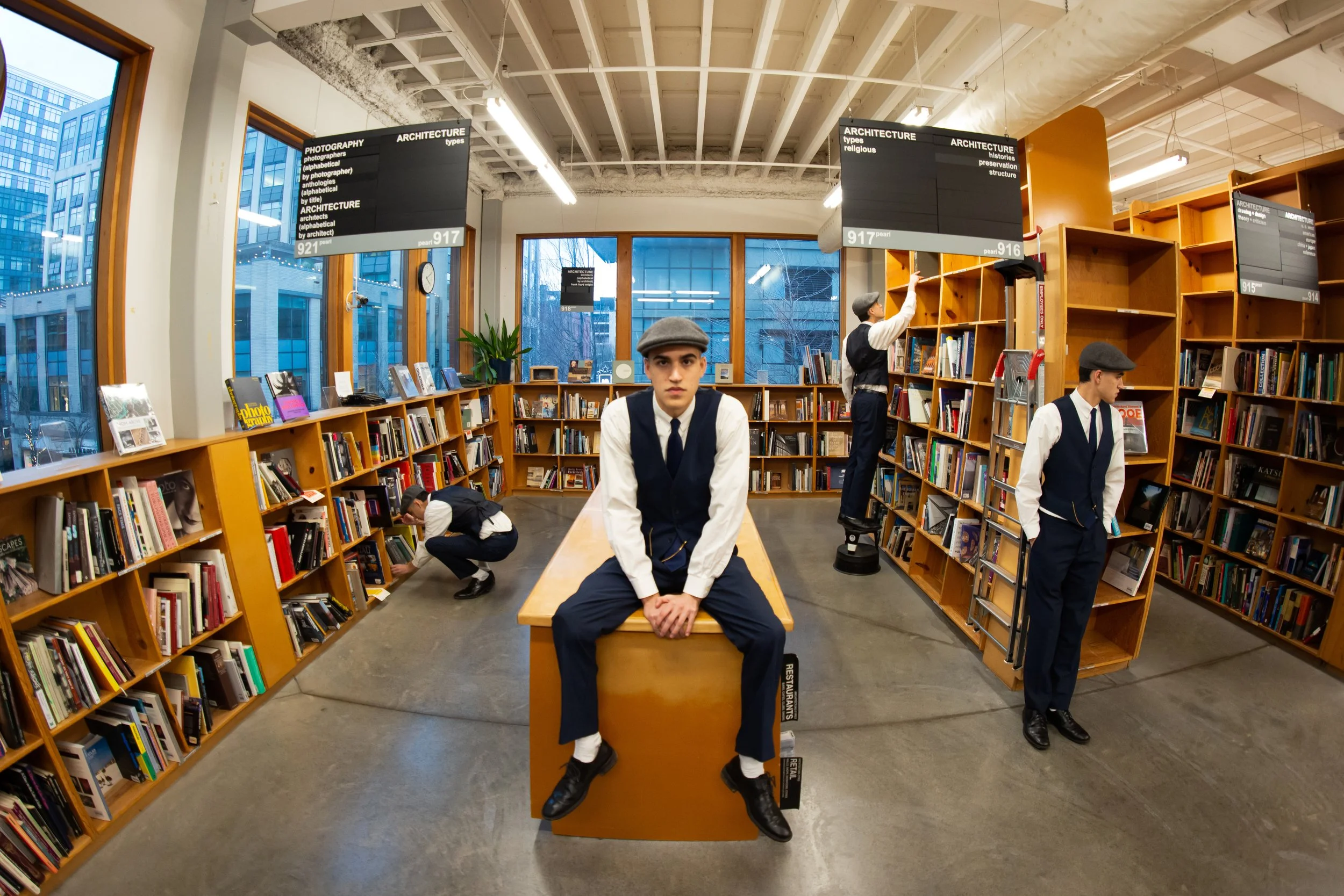 People dressed in vintage clothing inside a bookstore with wooden shelves, large windows showing city buildings, and digital screens hanging from the ceiling displaying information about architecture and photography.