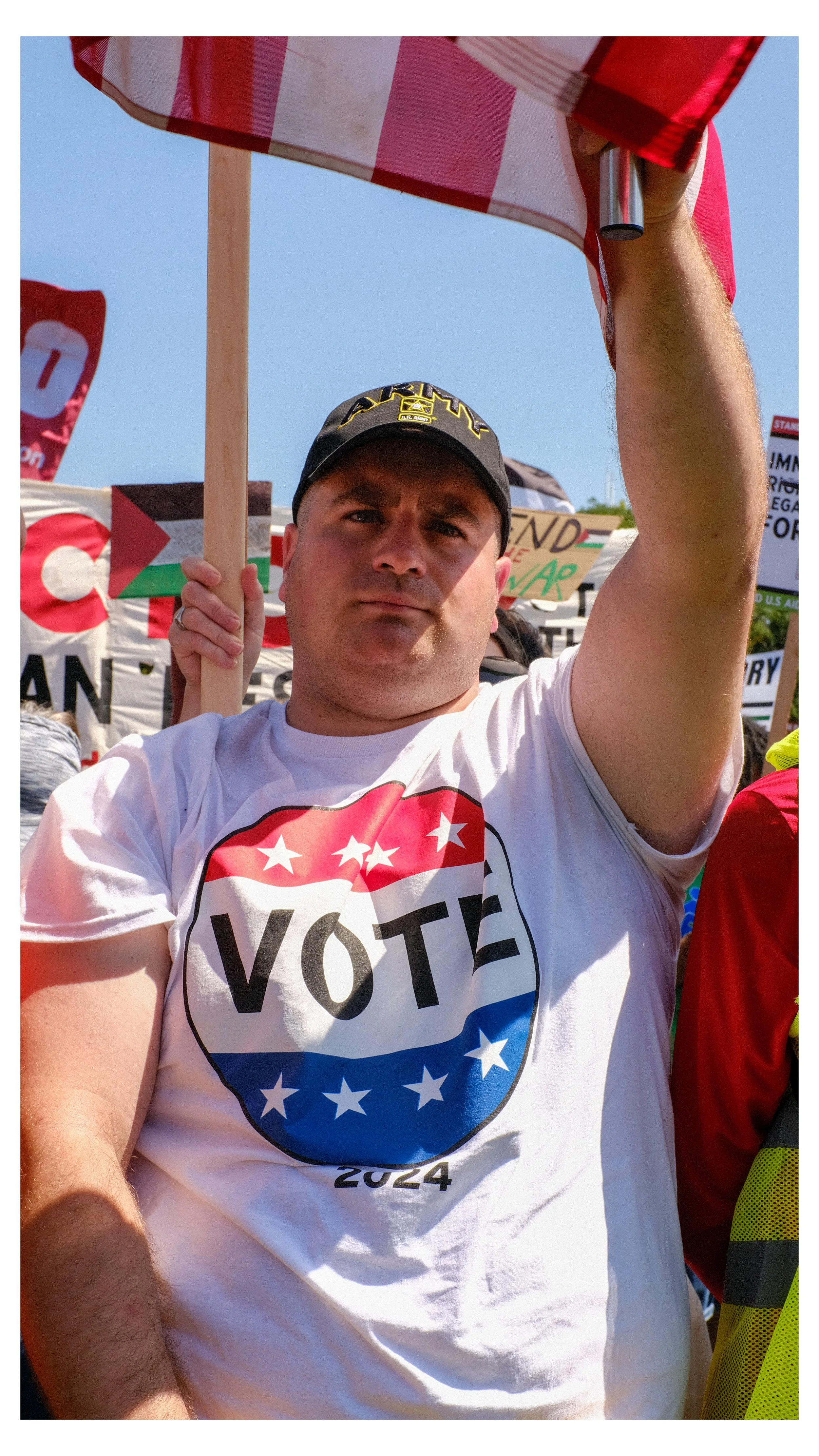 A man wearing a black baseball cap and a white T-shirt with a patriotic 'VOTE 2024' logo stands at a protest holding a red and white striped flag. Several signs and flags are visible in the background.