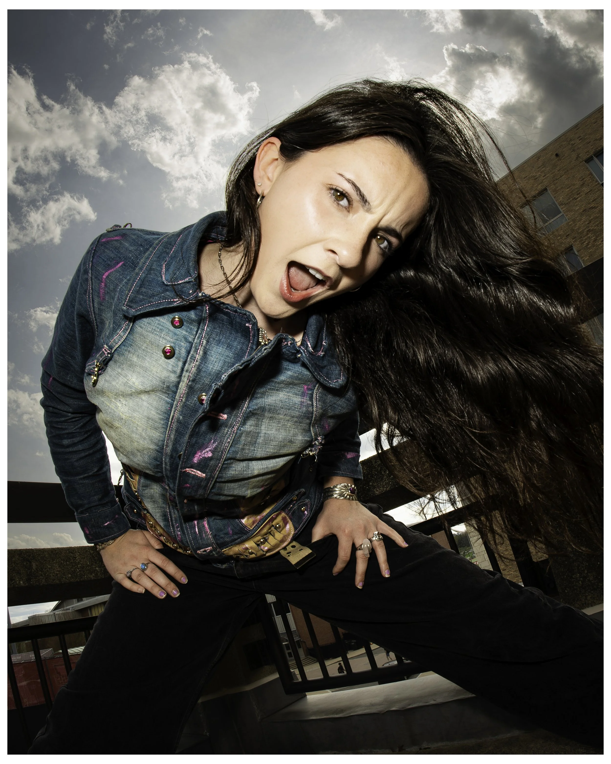 A woman with long dark hair is leaning forward outdoors with arms akimbo, showing her midsection. She is wearing a denim jacket with colorful stitching and accessories including rings, earrings, a chain necklace, and a decorative belt. The sky behind