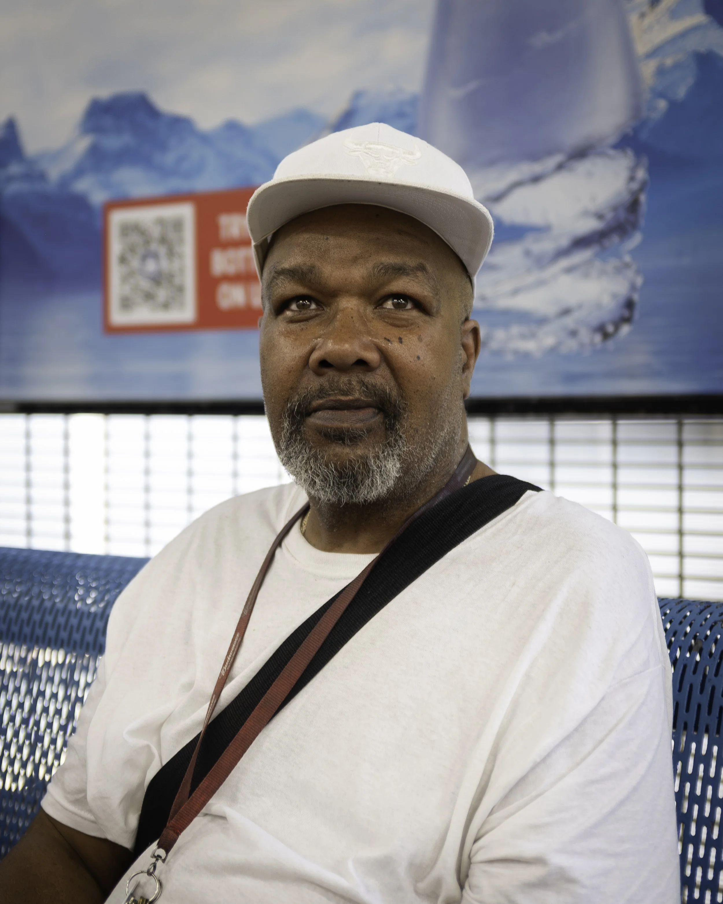 A man wearing a white baseball cap and white shirt, with a camera strap around his neck, sitting on a blue perforated metal bench with a mountainous background and a QR code on a sign behind him.