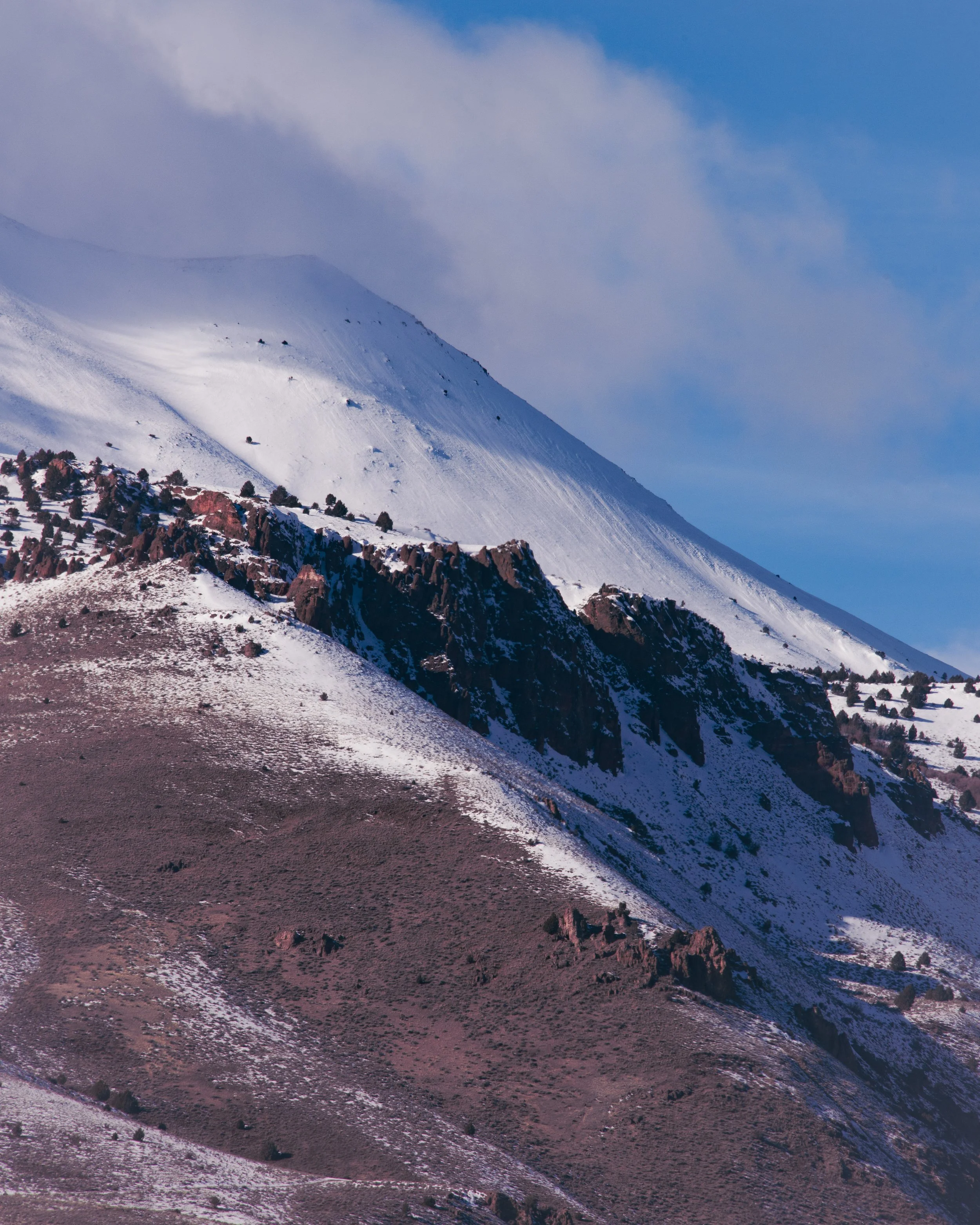 Snow-covered mountain slopes with rocky outcroppings and sparse trees, partially cloudy sky above.