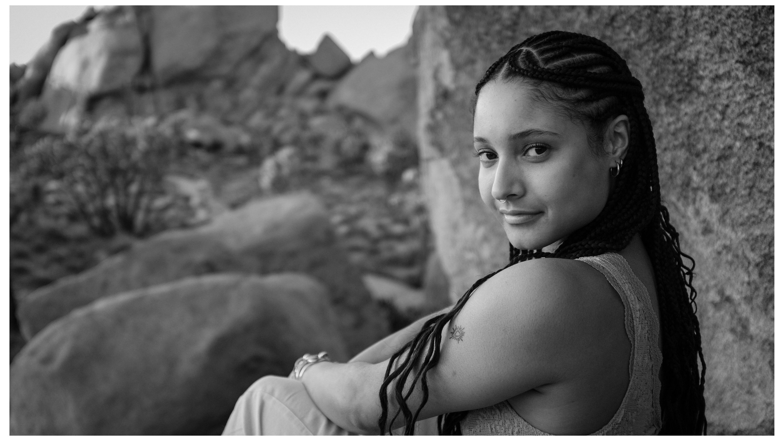 A young woman with braided hair and tattoos, smiling and looking over her shoulder near a rocky outdoor setting in black and white.