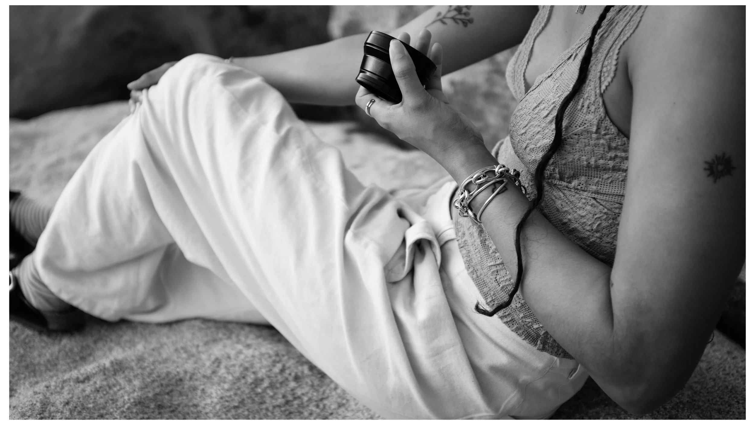 Black and white photo of a person lying on the ground, holding a small black camera in their right hand. The person is wearing a sleeveless top, jewelry, and has tattoos on their arm. Their legs are crossed, and the background appears to be grass or 