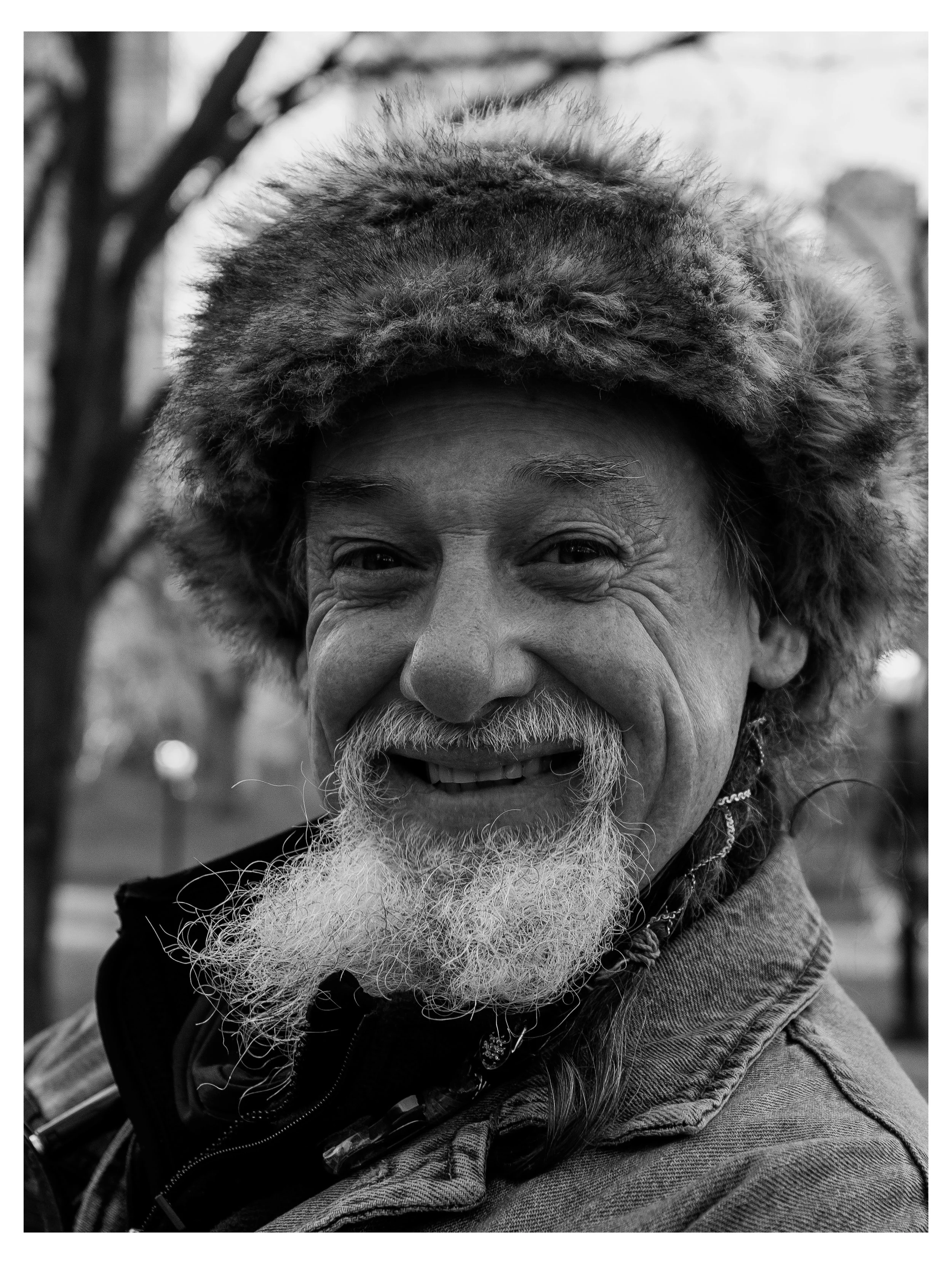 Close-up black and white photo of an elderly man smiling, wearing a fur-lined hat and a denim jacket.