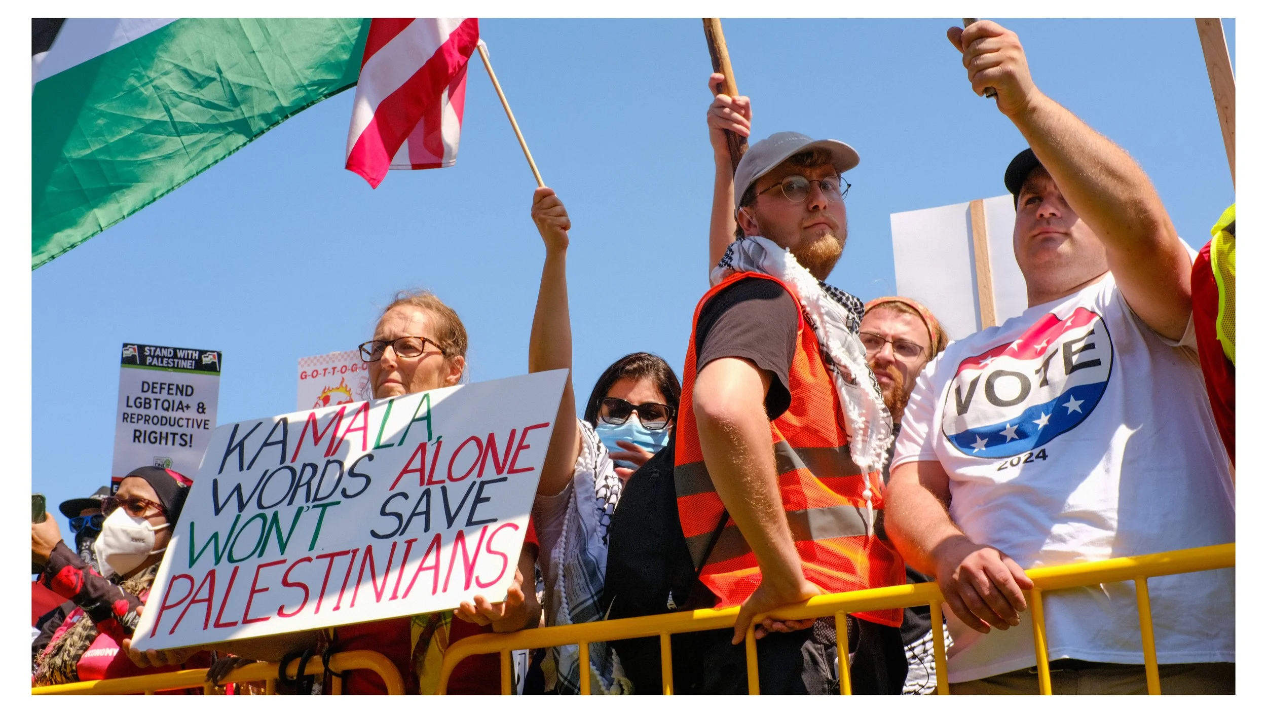 Group of people at a demonstration or rally holding signs and flags, including the Palestinian flag, under a clear blue sky.