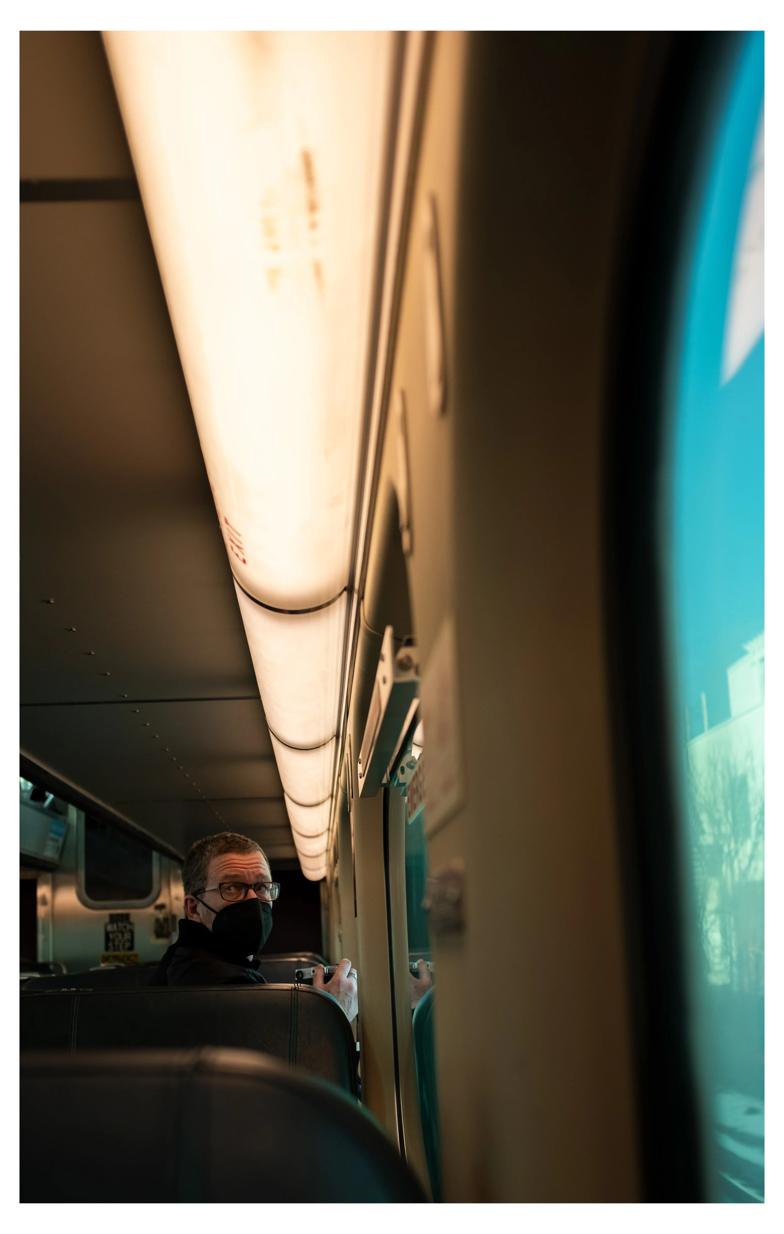 A man with glasses and a face mask sitting on a bus, holding a smartphone, with overhead bus lighting and window reflections.