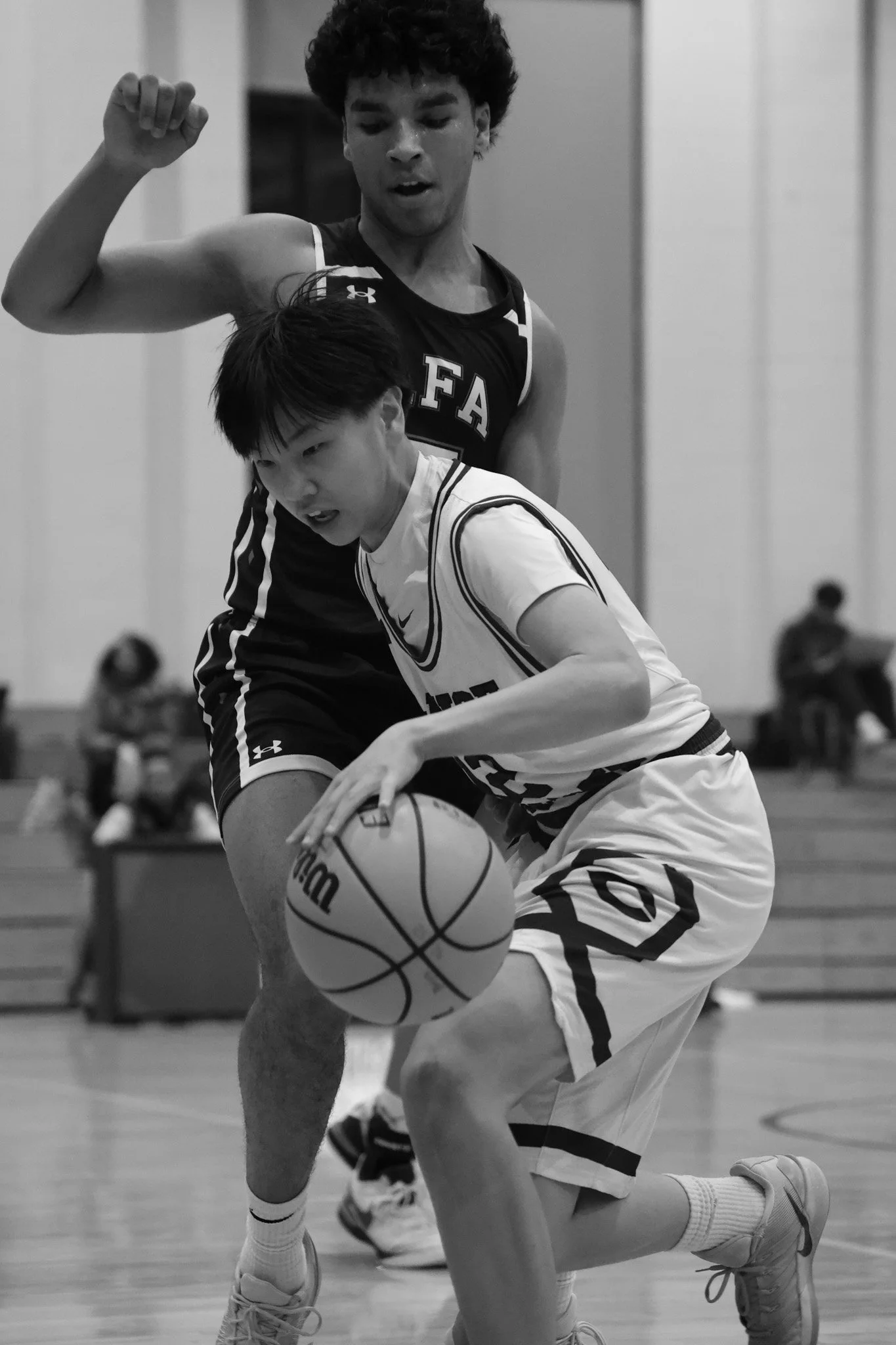 Two basketball players are competing in a game. The player in black is jumping, while the player in white is on the ground with one knee down, reaching for the basketball. The scene appears to be in an indoor gym with a blurred background.