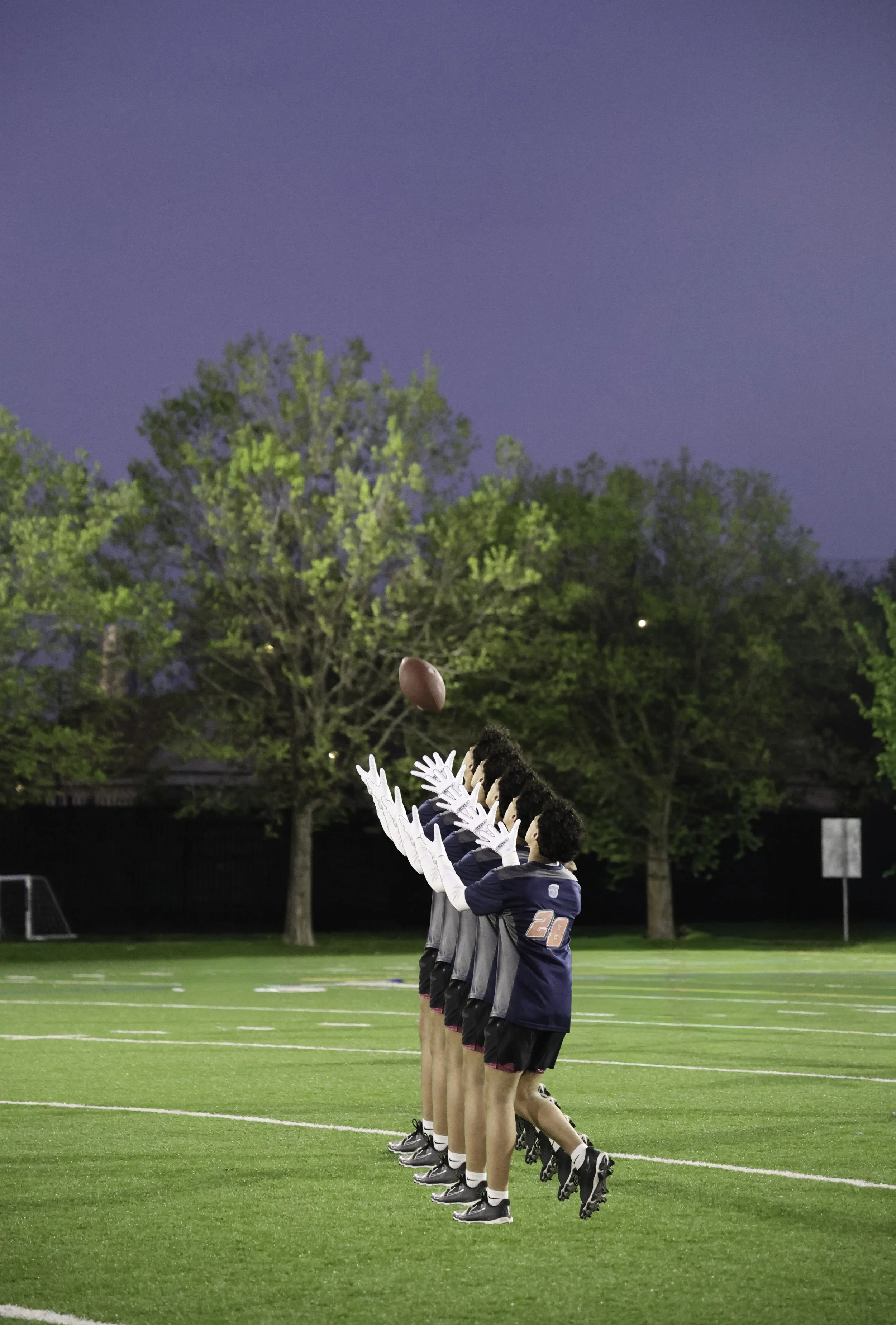 A group of athletes standing in a line on a football field during dusk, holding footballs above their heads, wearing uniforms and cleats, with trees and goalposts in the background.