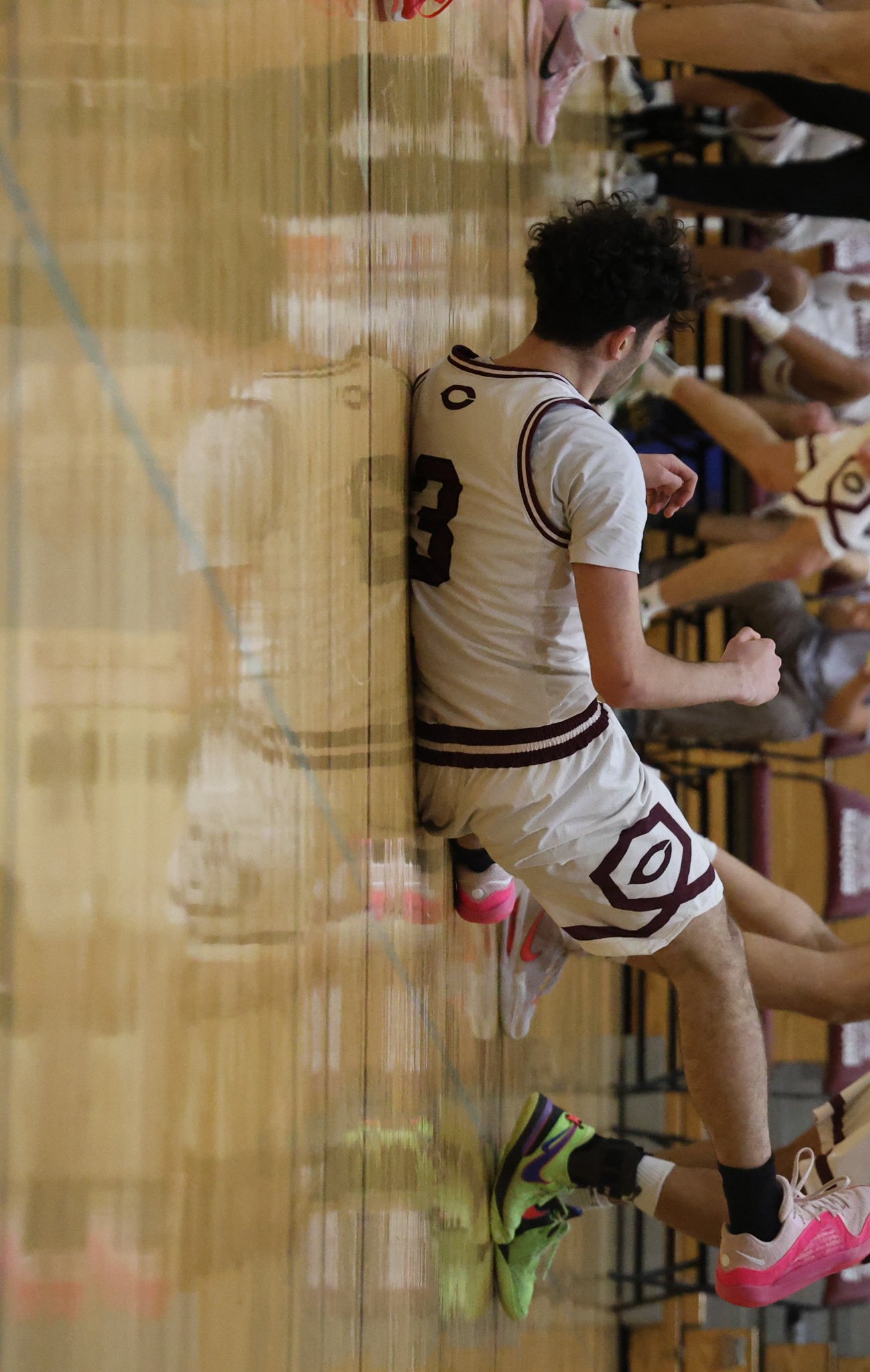 A person sitting on the basketball court floor leaning against the wall, wearing a white jersey and shorts, with colorful sneakers and socks. Other players are also sitting on the floor nearby.
