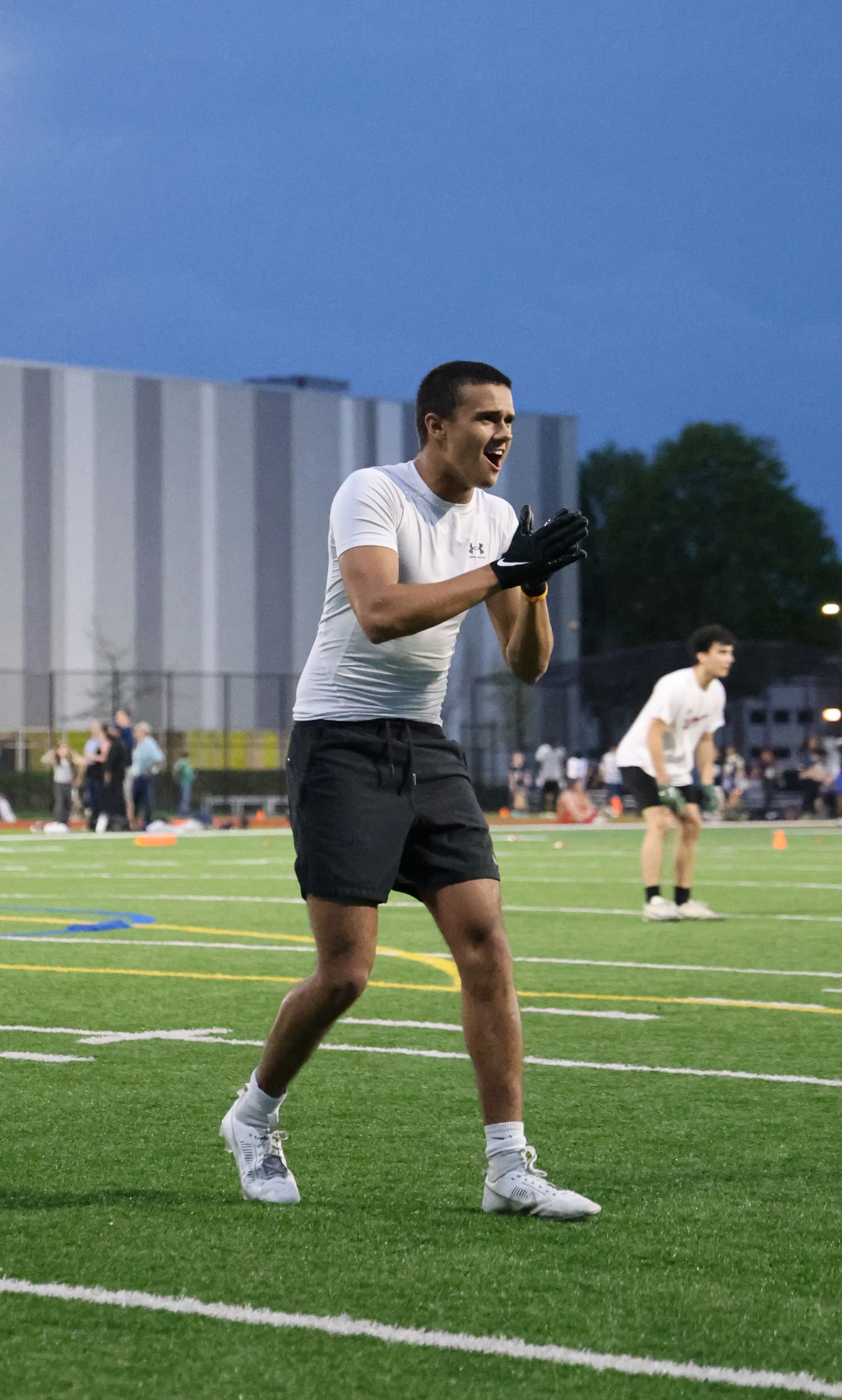 A young male football player in a white shirt, black shorts, and white sneakers on a football field during twilight, appears to be clapping or celebrating, with other players and spectators in the background.