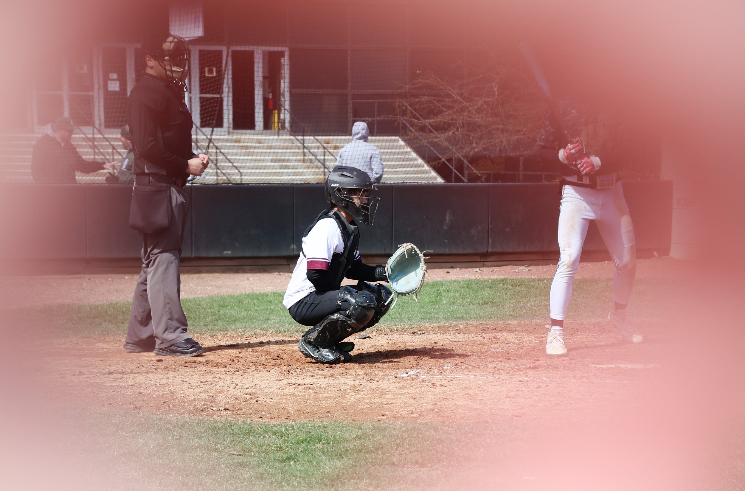A baseball catcher crouching behind home plate with a glove, a batter standing with a bat, a pitcher at the mound, and an umpire standing nearby, on a baseball field during daytime.