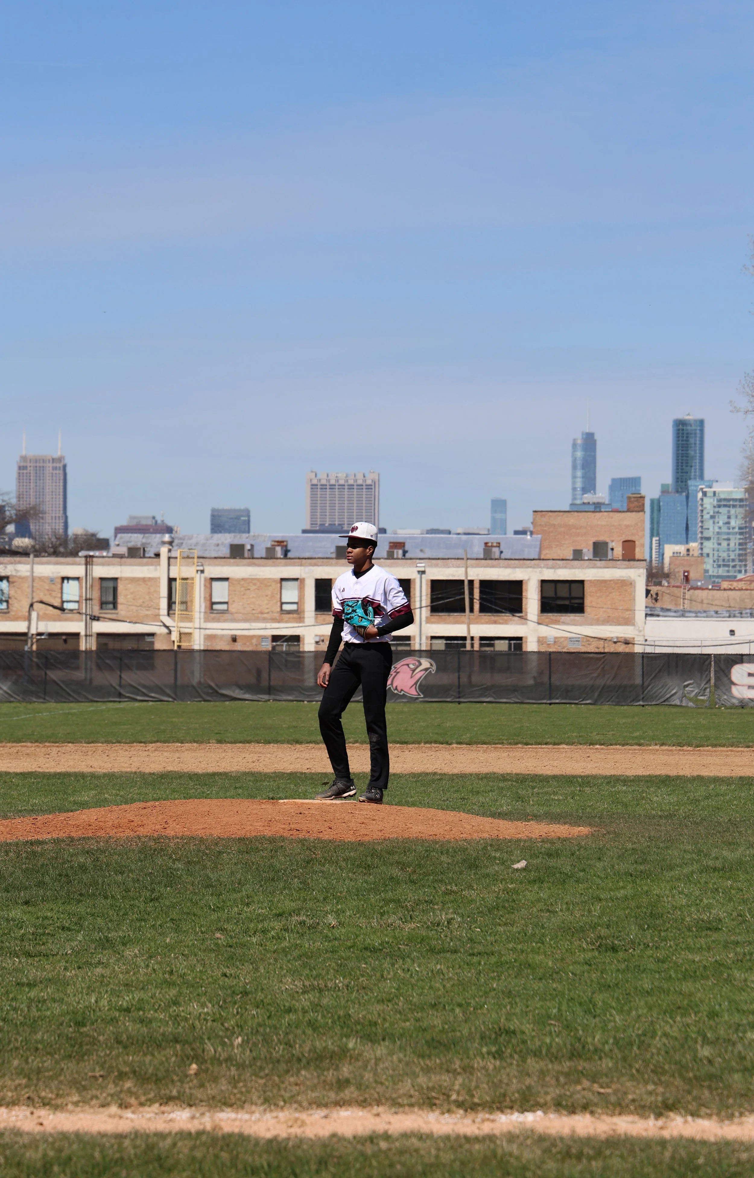 A baseball player standing on the pitcher's mound on a baseball field with a city skyline in the background.