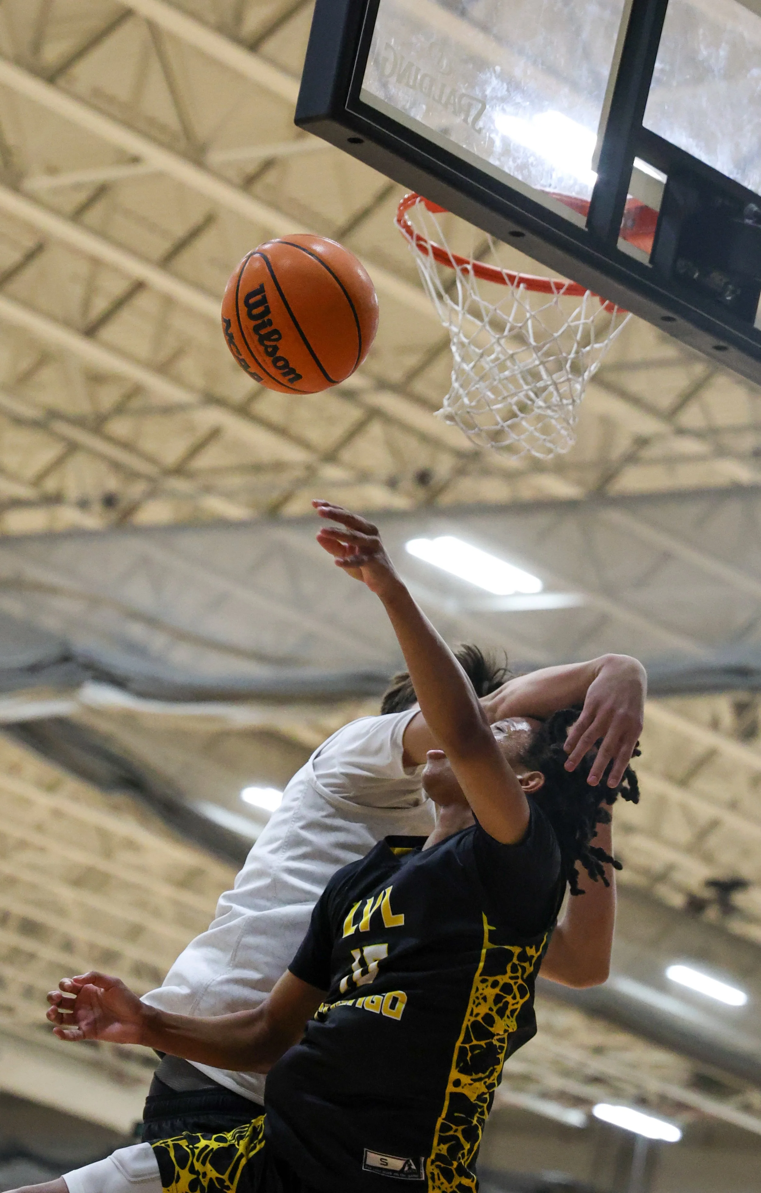 A basketball game in progress with players jumping near the hoop to contest a shot, with the basketball in mid-air heading toward the basket.