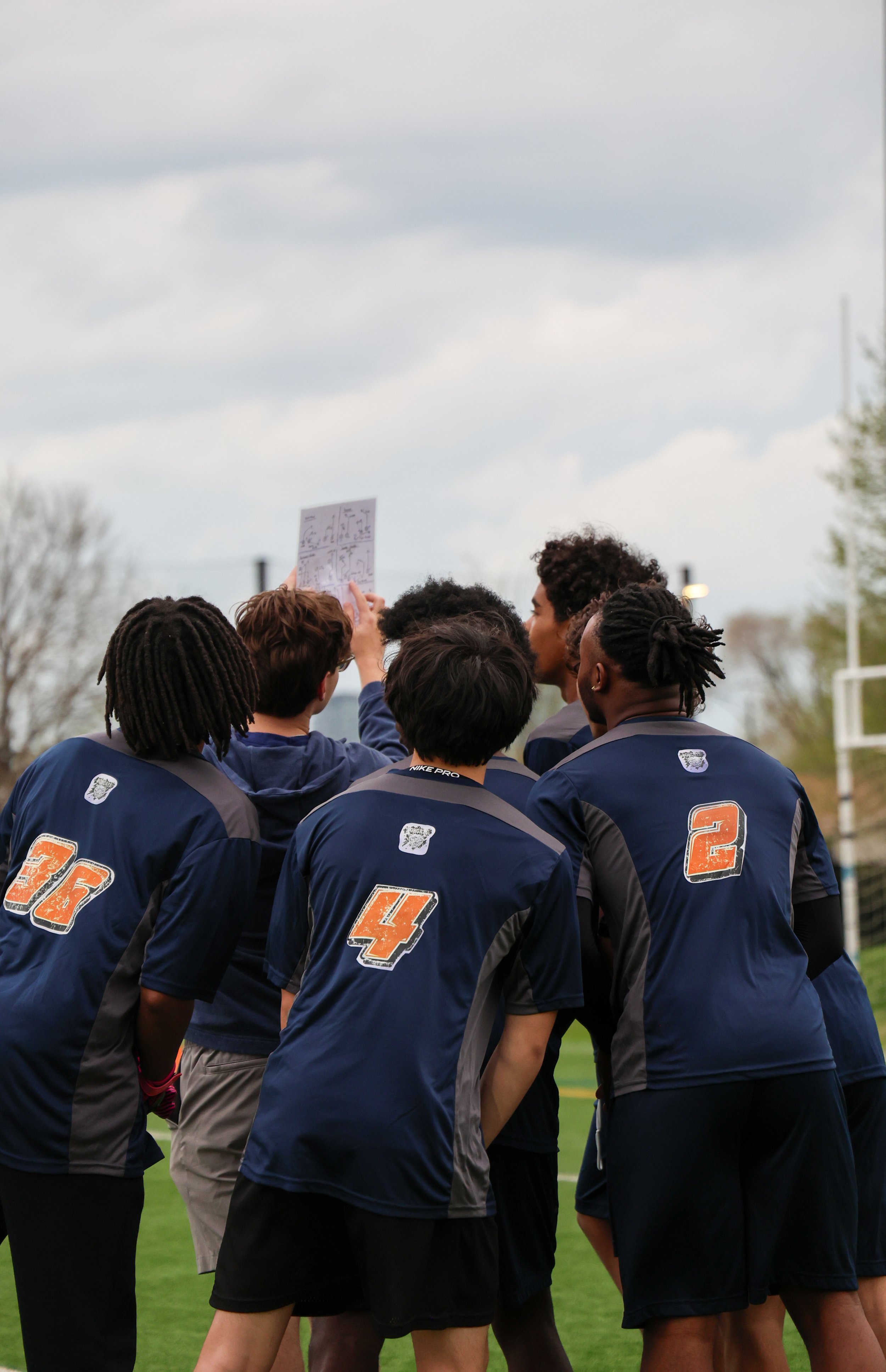 A group of young athletes in navy blue sports jerseys with numbers on the back, standing outdoors on a field, gathered in a huddle. One person is holding a white sheet with diagrams or notes. The sky is cloudy, and there are trees and a football goalpost in the background.