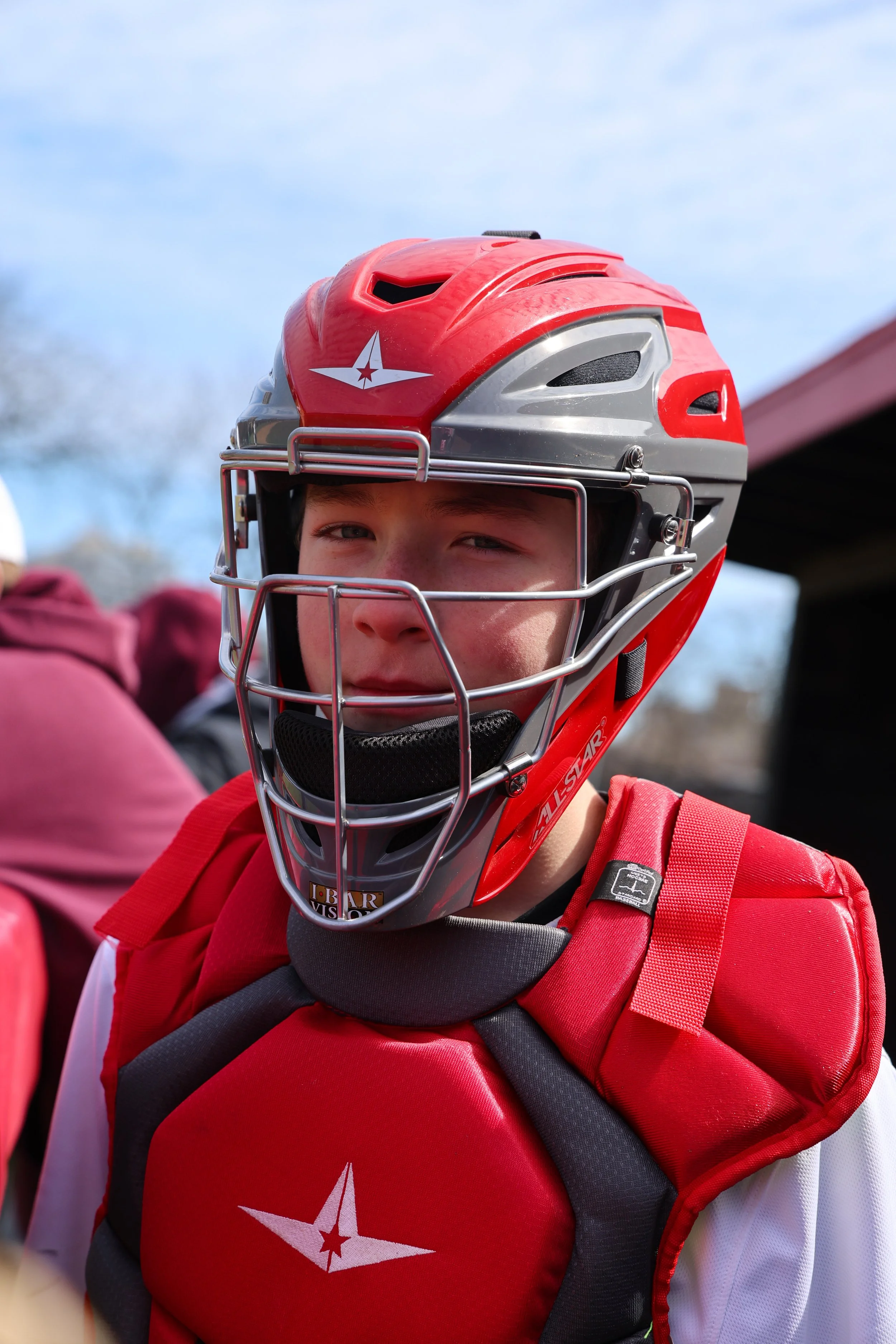 A young ice hockey player wearing a red and gray helmet with a face cage and a red jersey with a star logo, carrying a black backpack, outdoors on a sunny day.