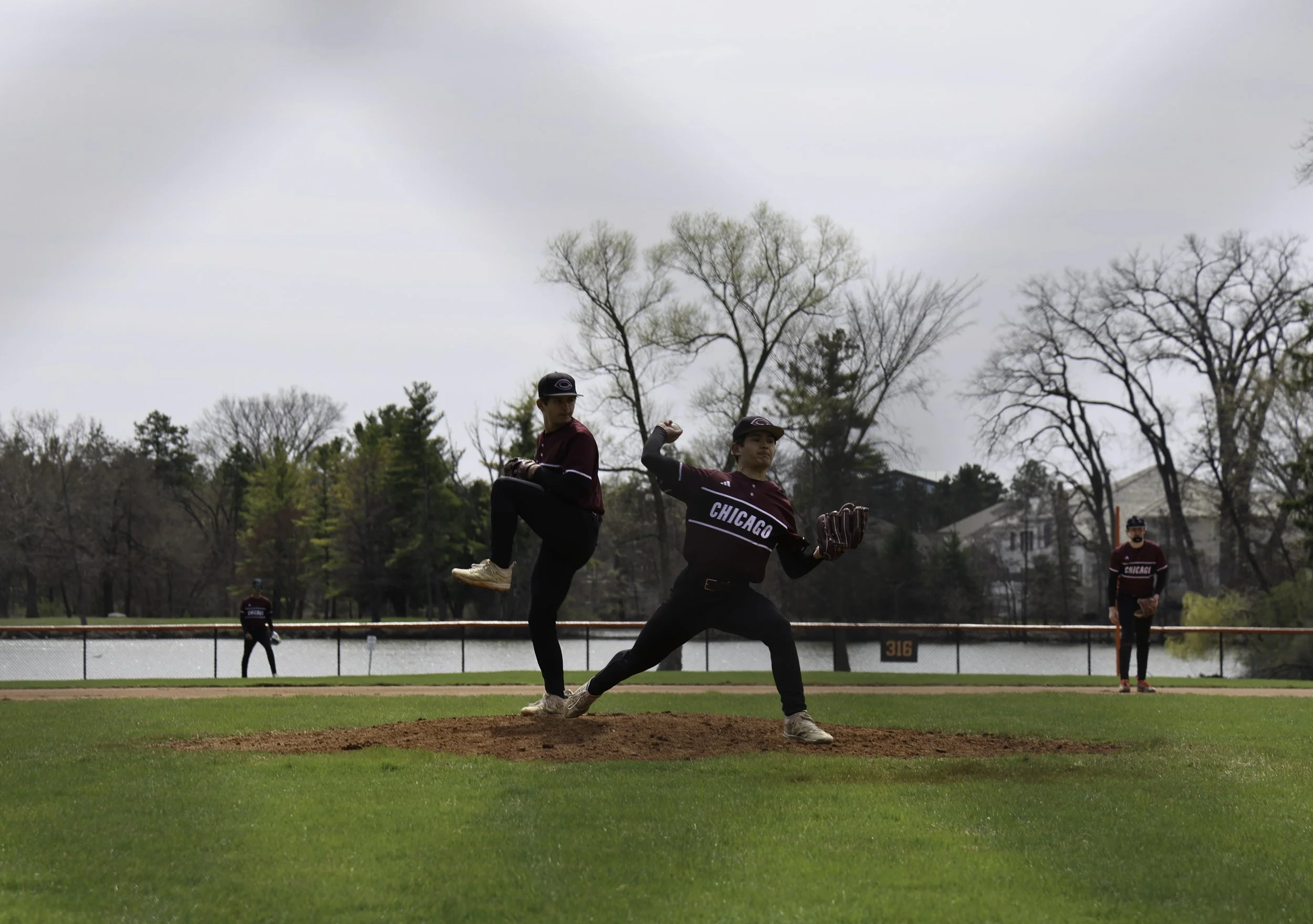 Two young men in Chicago baseball uniforms practicing pitchers' mound throws on a baseball field during daytime with overcast skies, trees, and houses in the background.
