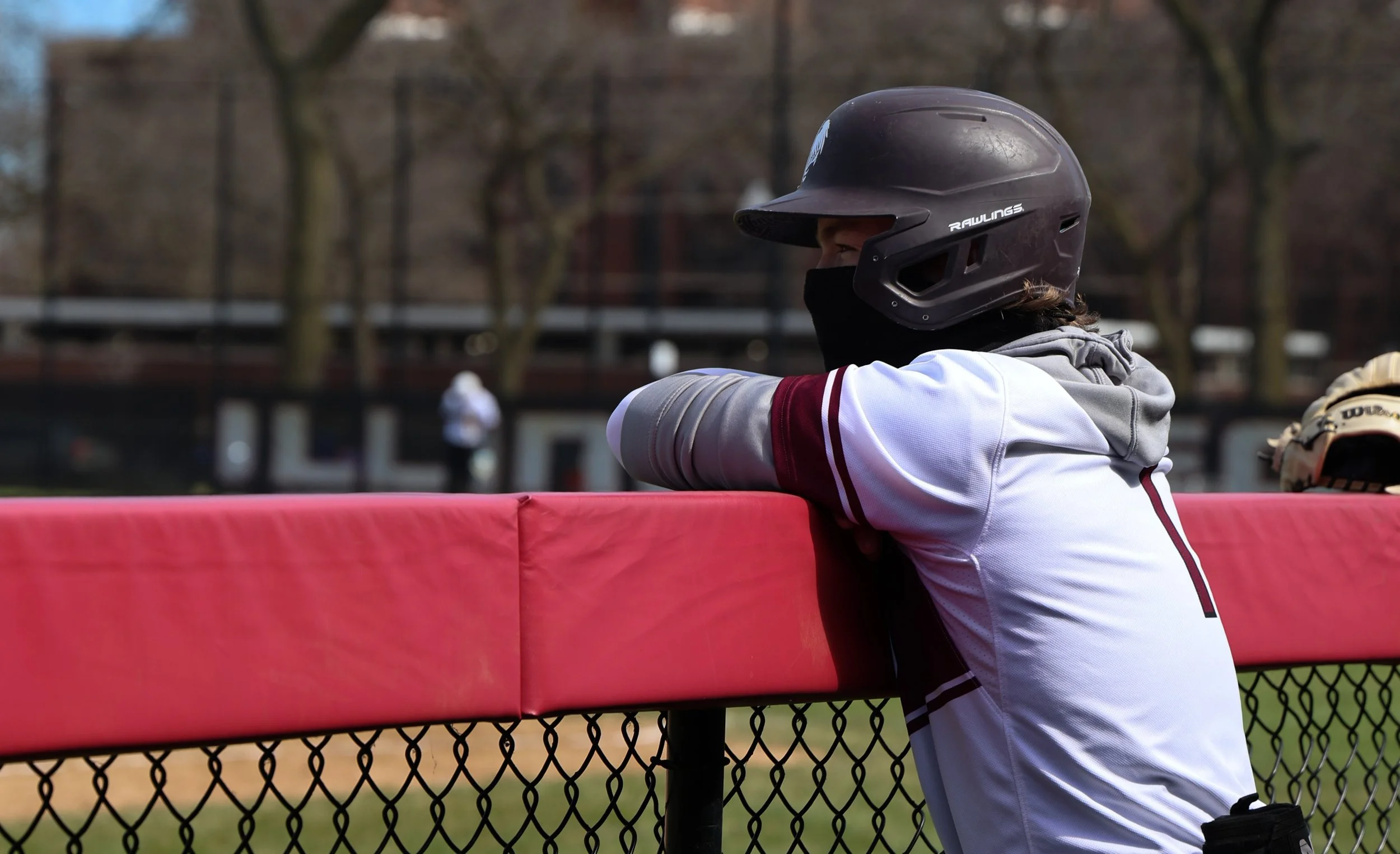A person wearing a black helmet and a face covering, leaning on a fence at a baseball or softball field, with trees and a chain-link fence in the background.