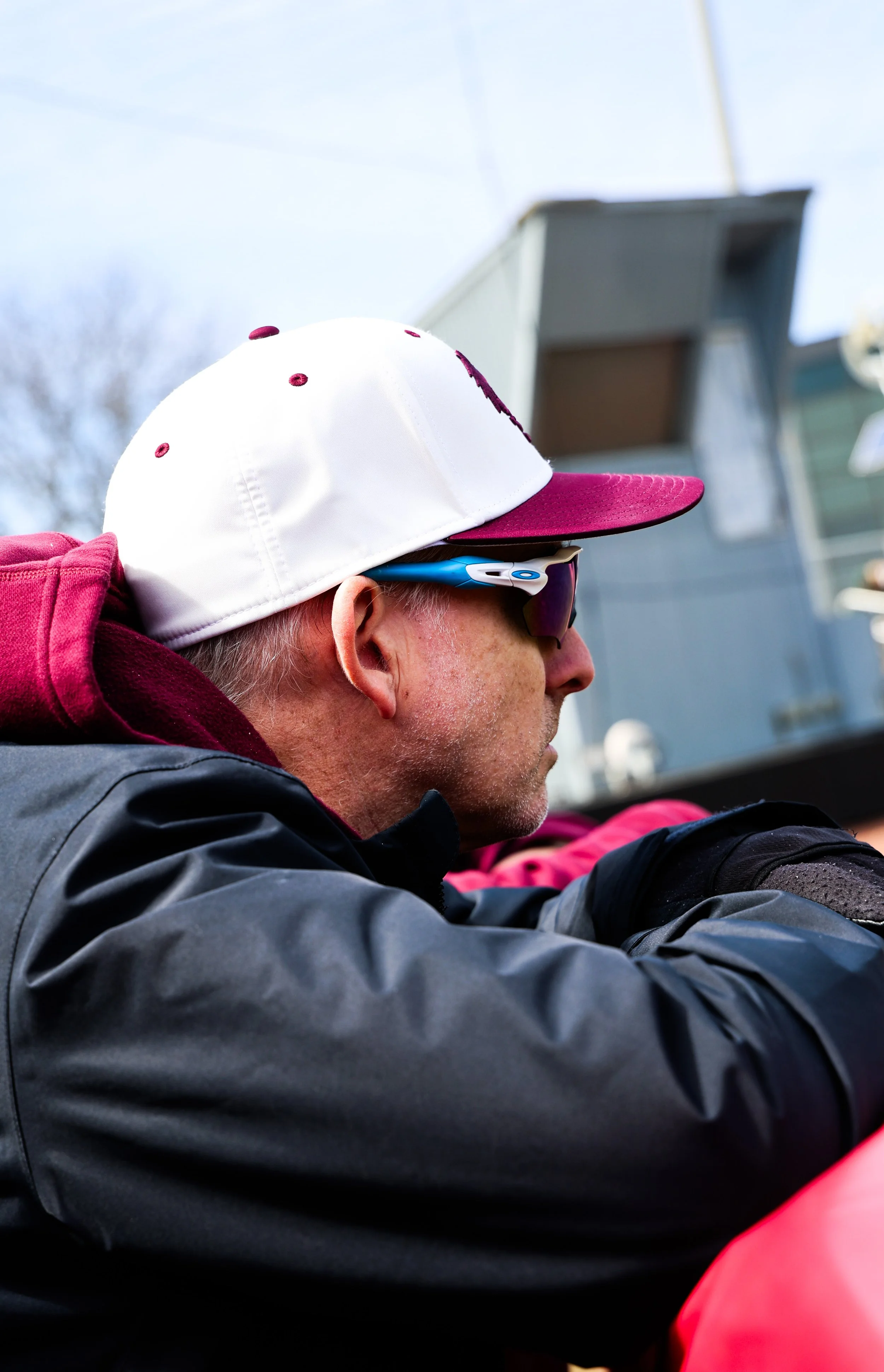 Close-up of a person wearing sunglasses, a white cap with a pink brim, and a maroon hoodie, outdoors with a blue sky and a building in the background.
