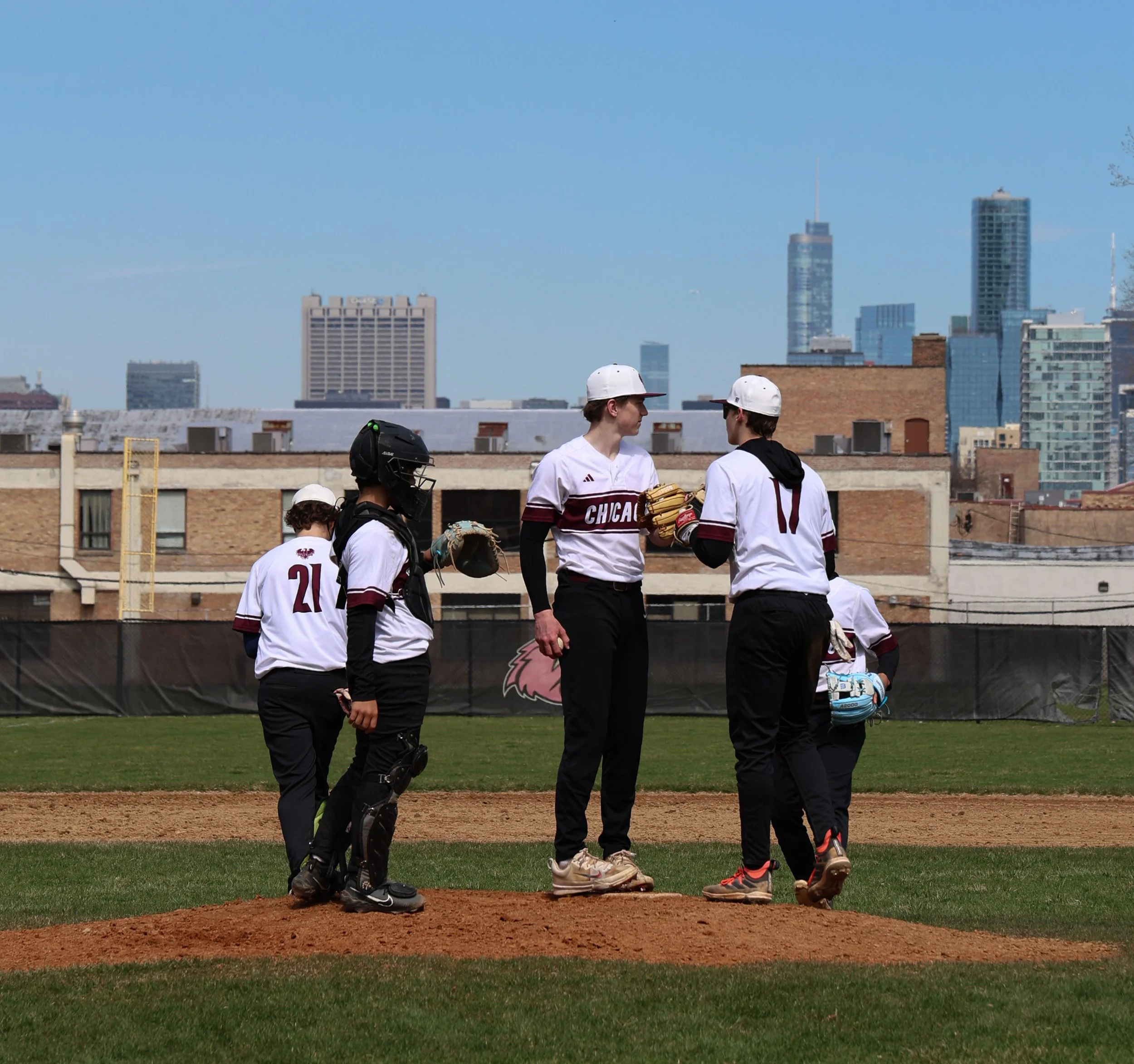 Young baseball players wearing uniforms and helmets standing on the field, with a city skyline in the background under a clear blue sky.
