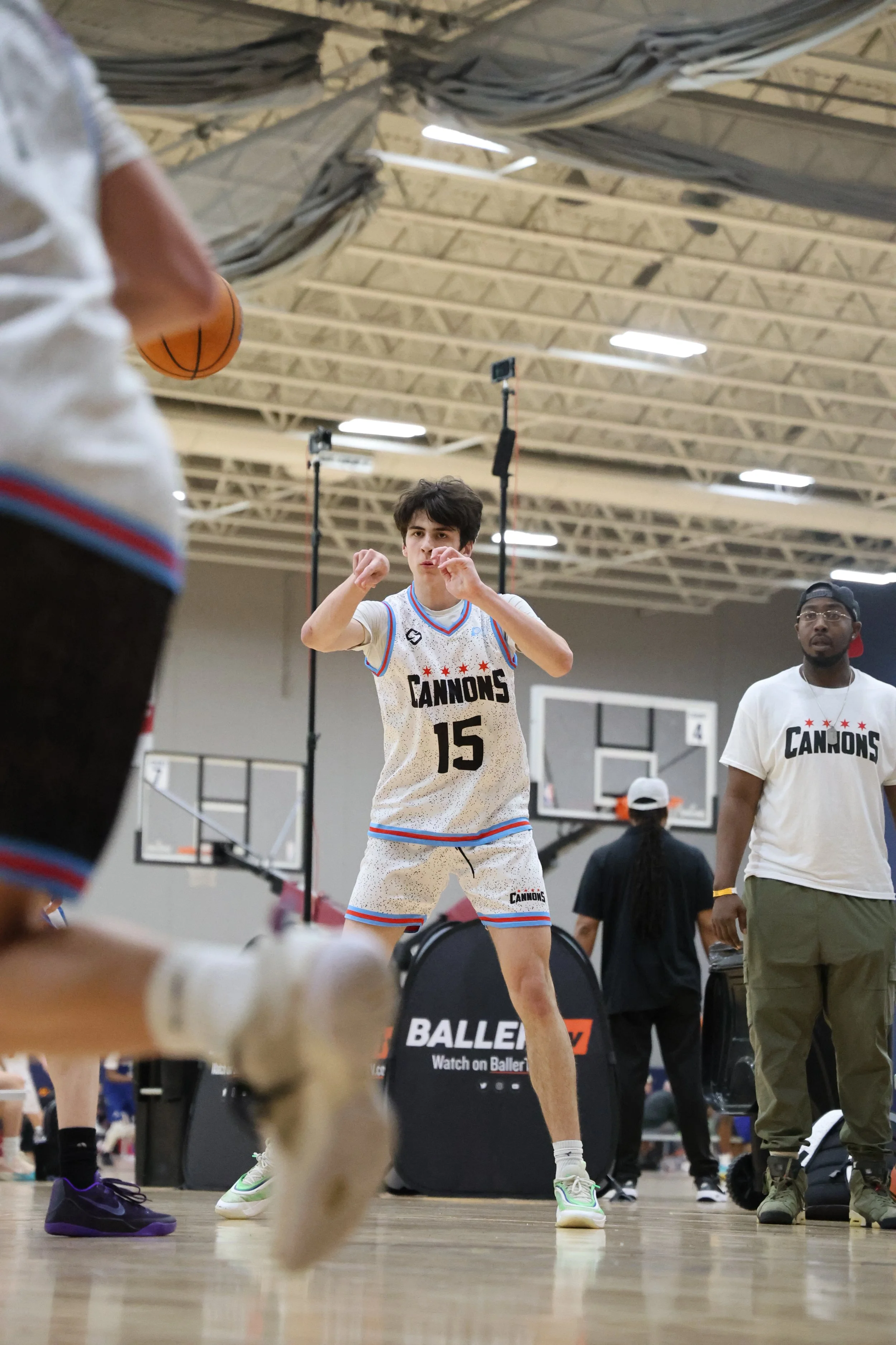 A basketball player wearing a white jersey with the number 15 and the team name 'CANNONS' is about to shoot the ball in an indoor gym. A person in casual clothing stands nearby, and other players and equipment are visible in the background.