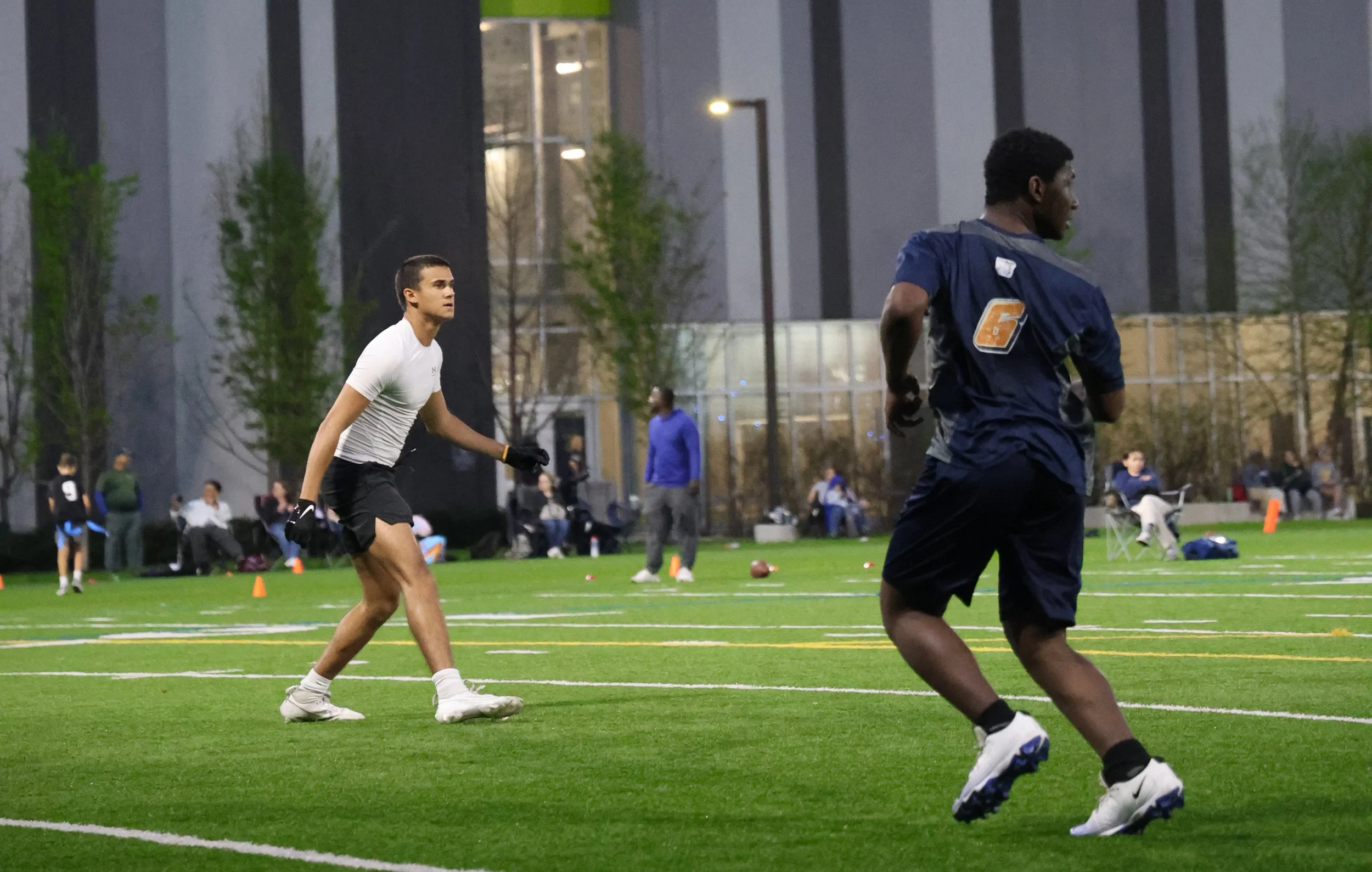 Two young men playing flag football on an outdoor field in the evening, with other players and spectators in the background.