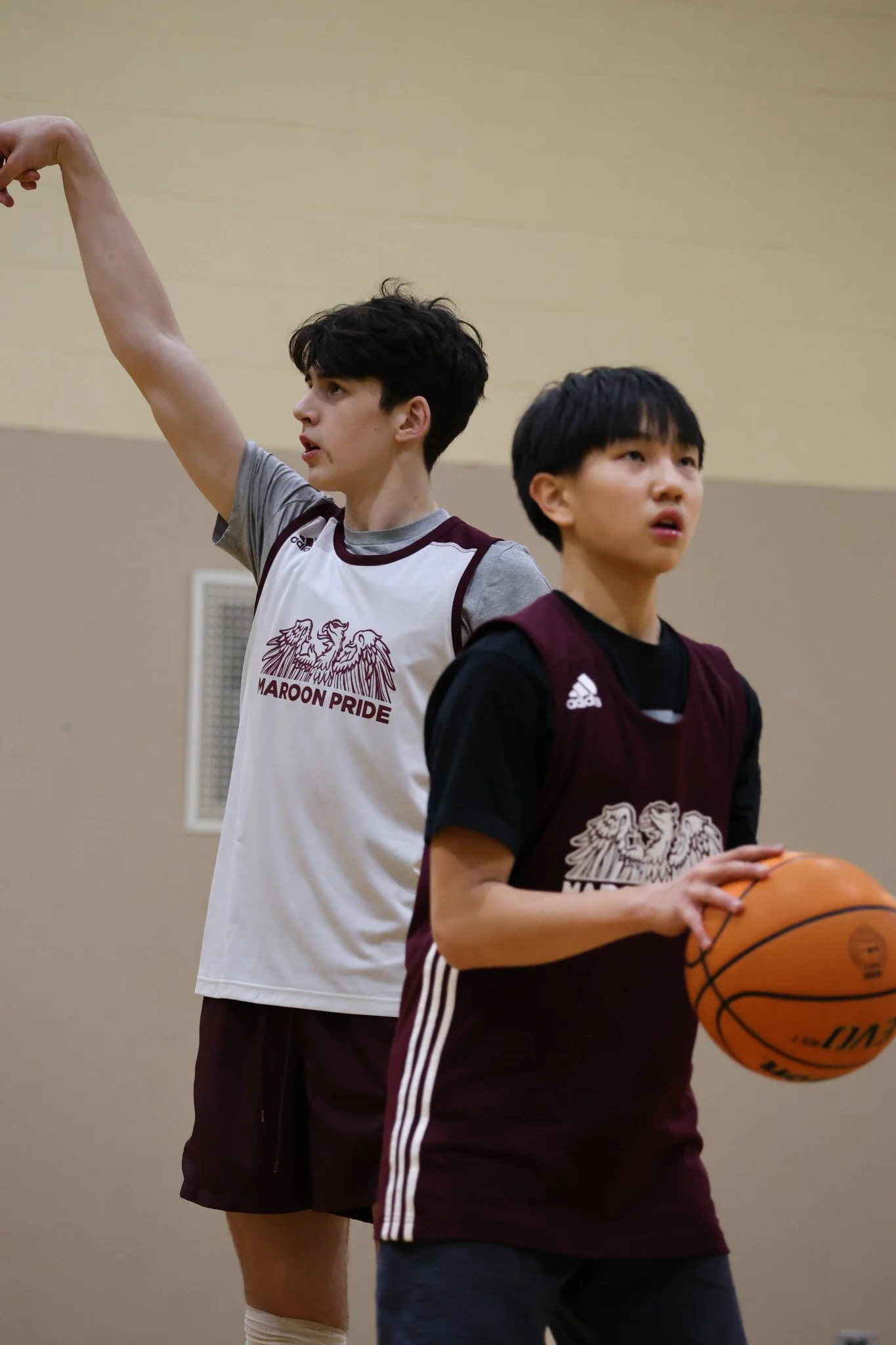 Two young males playing basketball indoors, one holding a basketball and the other raising his arm, both wearing Maroon Pride jerseys.