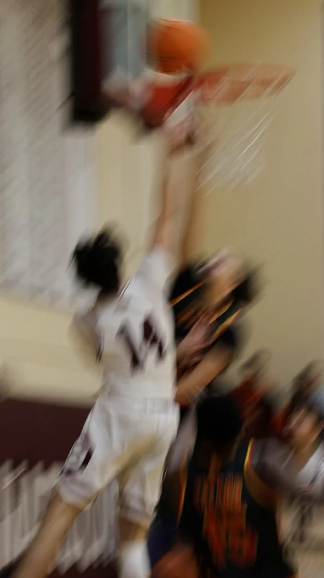 A blurred photo showing a basketball game with players in action, one attempting a shot at the hoop and others defending or moving around the court.