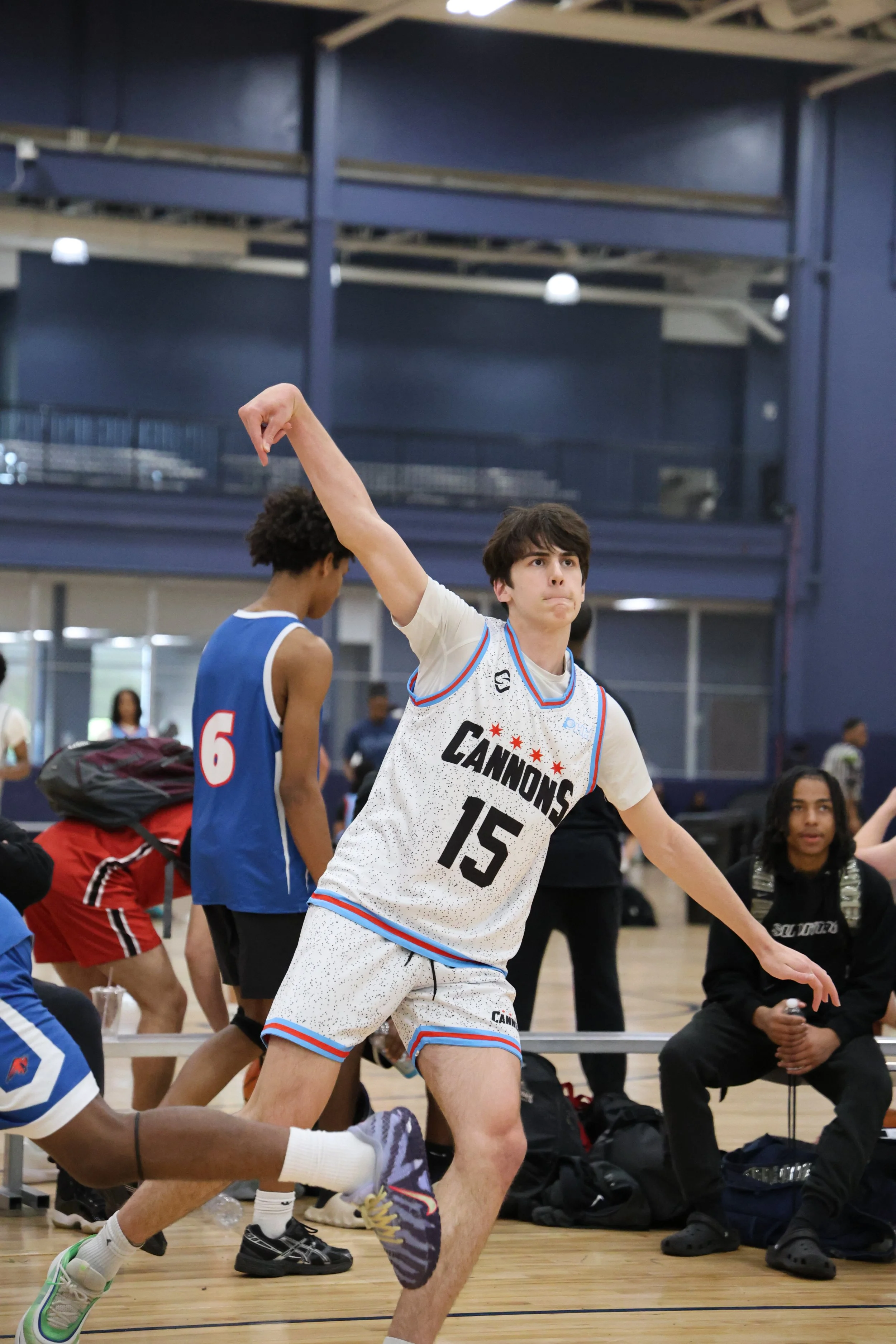 A basketball player in a white uniform with the words "CANNONS" and the number 15, preparing to shoot during a game or practice session at an indoor gym, with spectators and another player in the background.