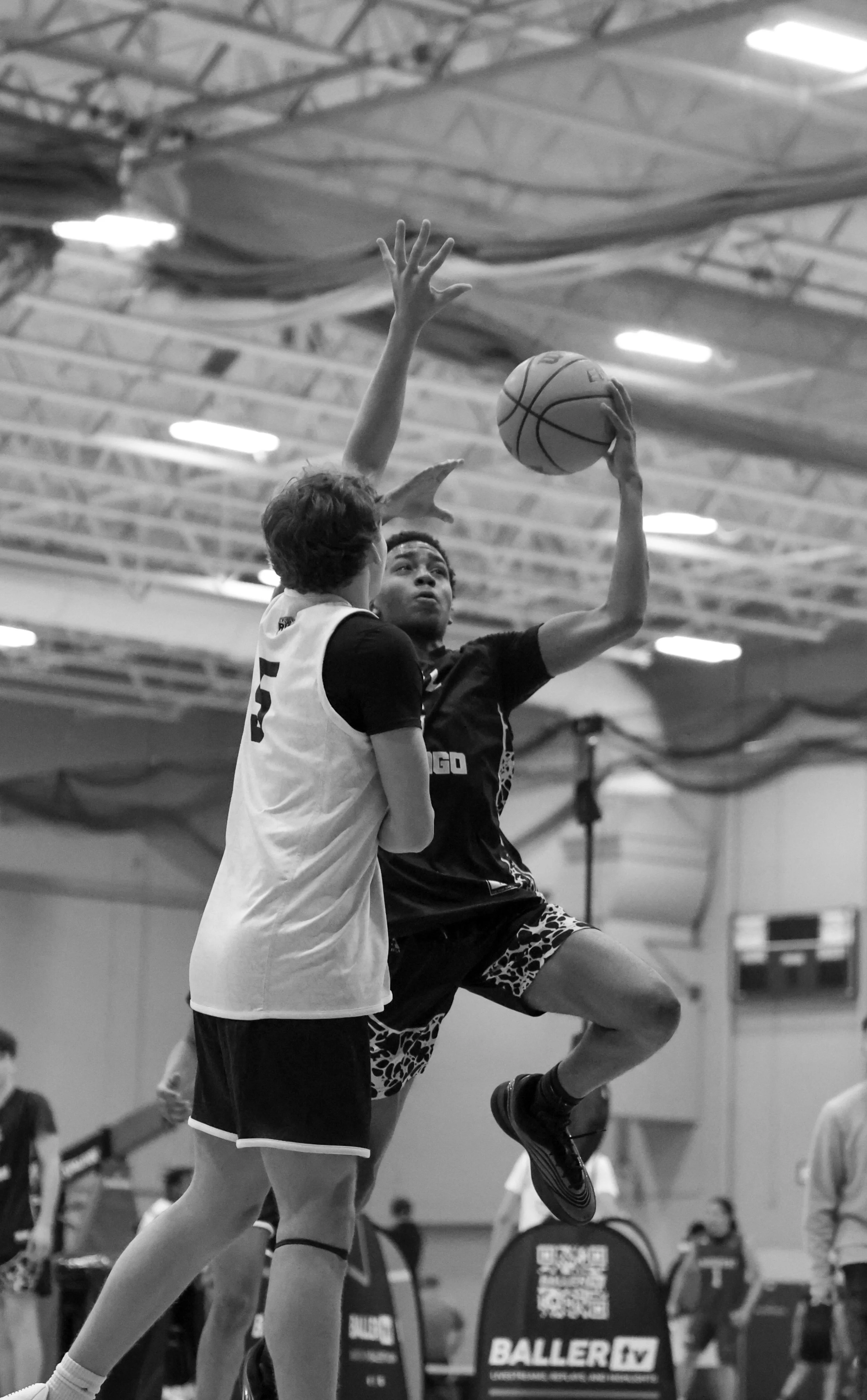 A woman in a sports uniform is jumping to shoot a basketball while a player in a white jersey is defending her in a gymnasium.