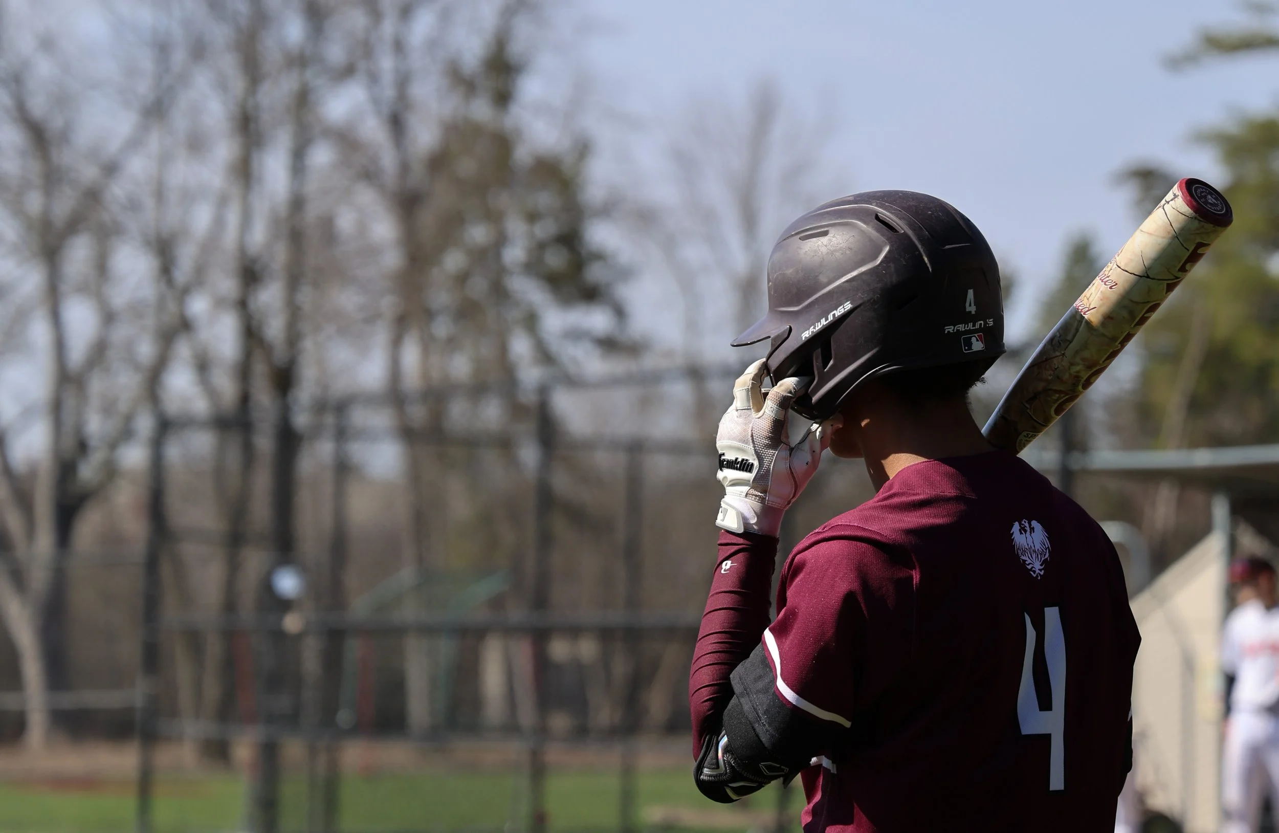 A baseball player wearing a black helmet and maroon jersey adjusts his helmet while holding a bat, with a baseball field and trees in the background.