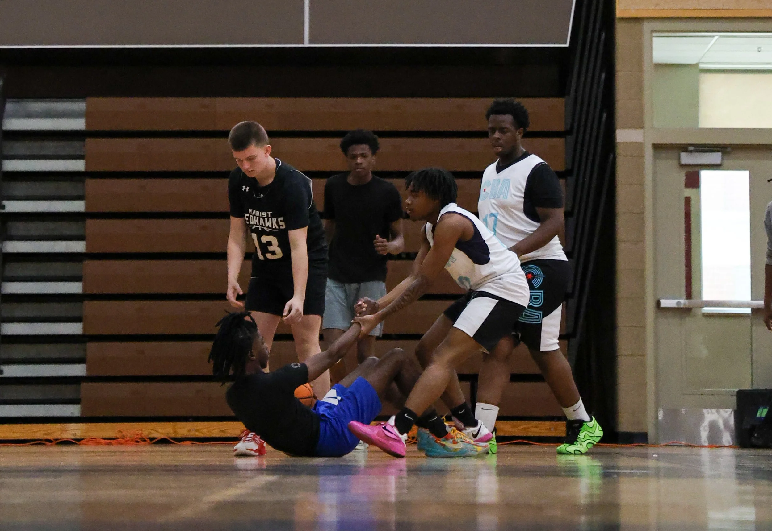 A group of young men playing basketball indoors, with one player on the floor reaching for the ball while others stand nearby.