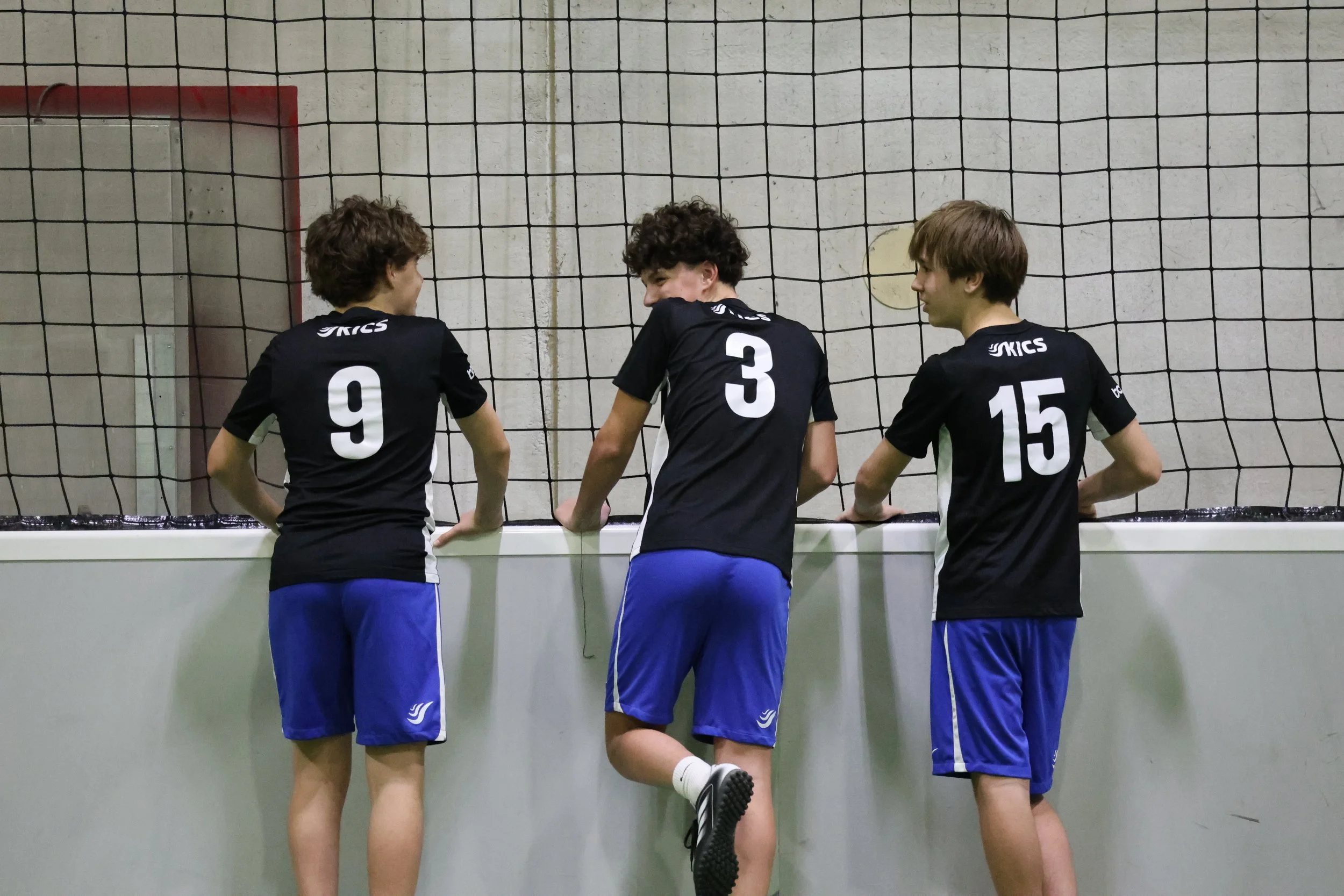 Three male athletes in black and blue sports uniforms standing in front of a volleyball net, facing away from the camera, inside a gymnasium.