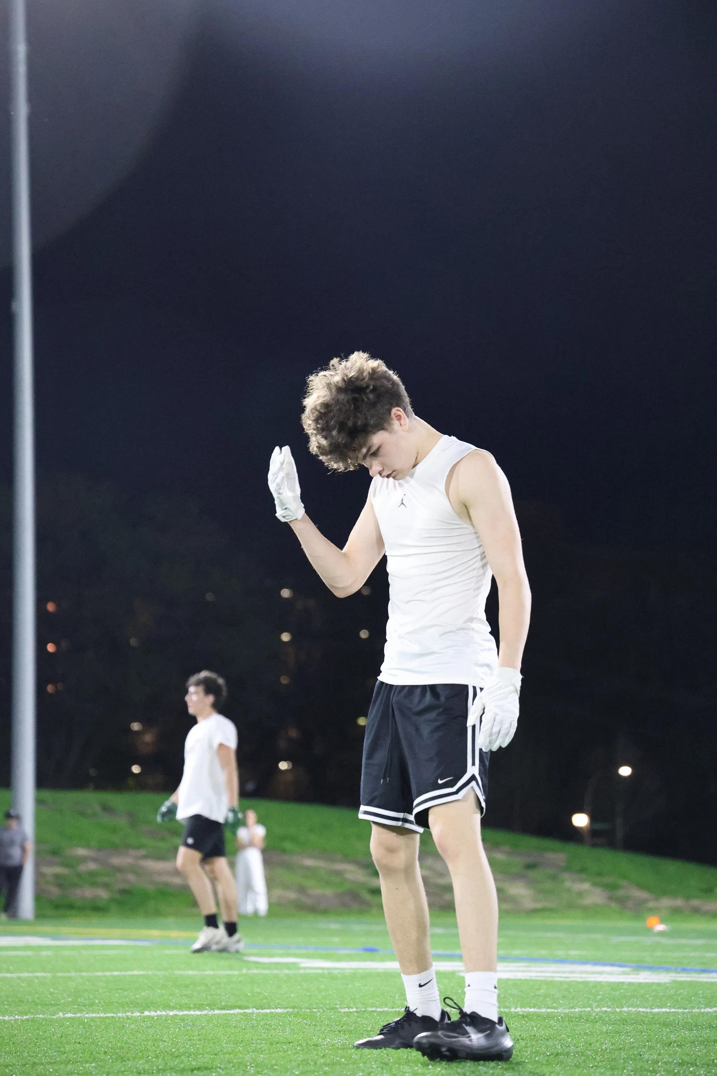 Young male athletes in white athletic clothing and gloves standing on a football field at night. One is in the foreground with his head bowed and hand raised, other athletes are in the background.