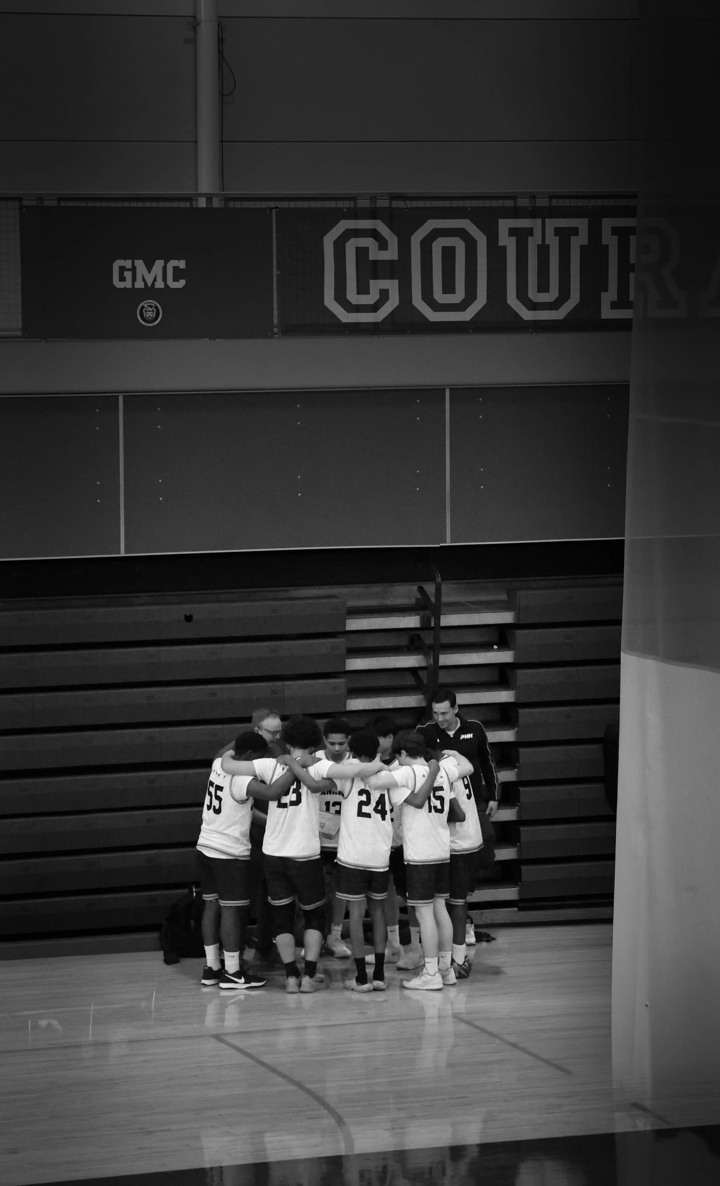 A group of basketball players in uniforms huddle together on the court during a game, with a coach standing nearby. The gymnasium has a wooden floor and seating in the background, with banners and a wall.