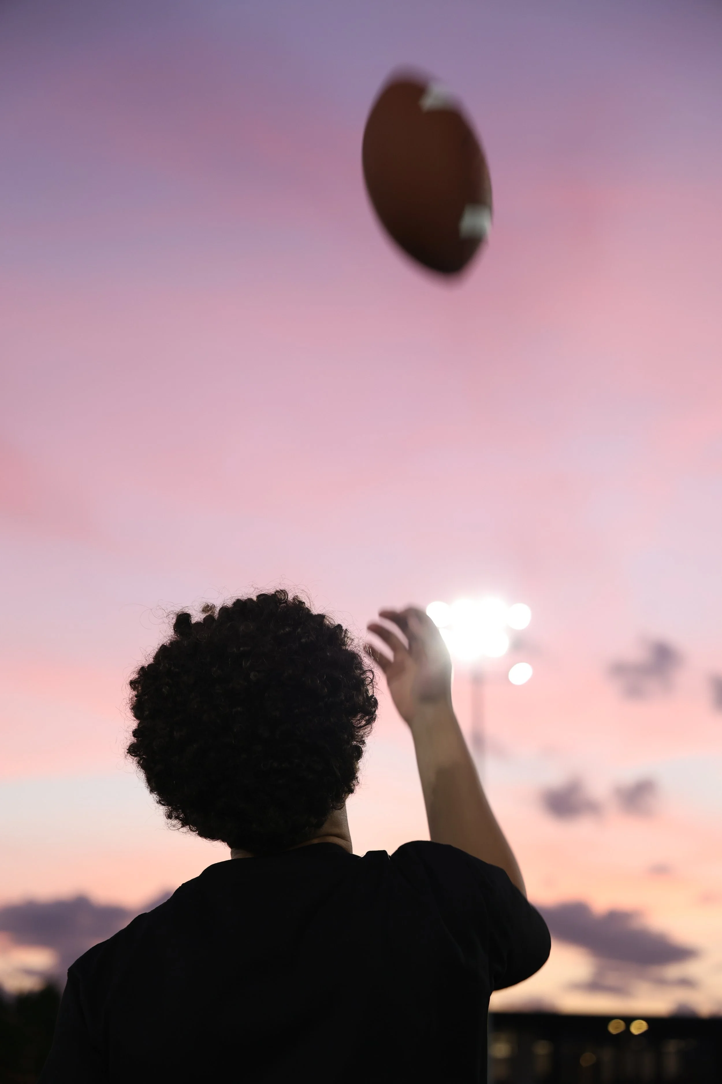 A person with curly hair is reaching up towards a floating hot air balloon during sunset, with a pink and purple sky in the background.