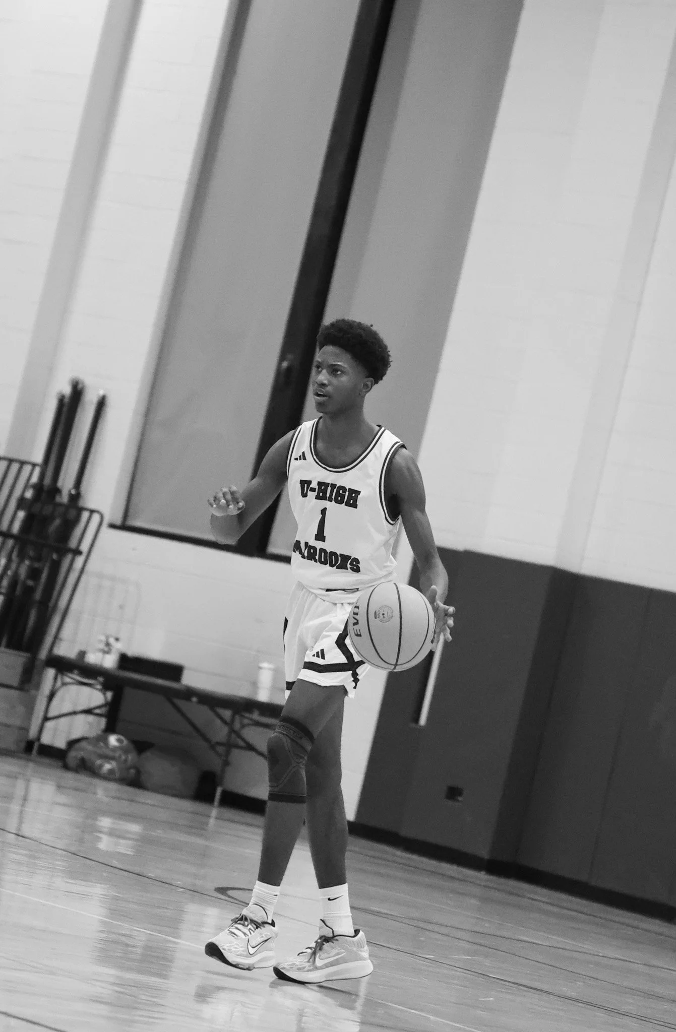 A young male basketball player dribbling a basketball on an indoor court, wearing a jersey with the logo 'V-USD Moore' and the number 1.